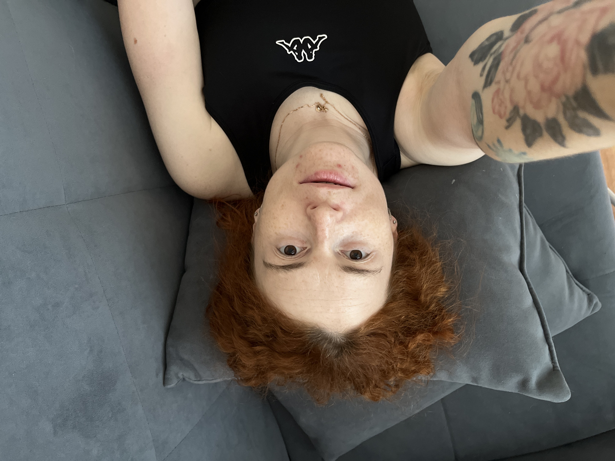 A person with red curly hair lies on a gray couch. The person is wearing a black tank top with a white Kappa logo. They are wearing a gold necklace. They have a tattoo on their right arm. The person's head rests on a gray pillow. The picture appears to be taken from a first-person perspective. The location is unspecified, therefore cannot be inferred.