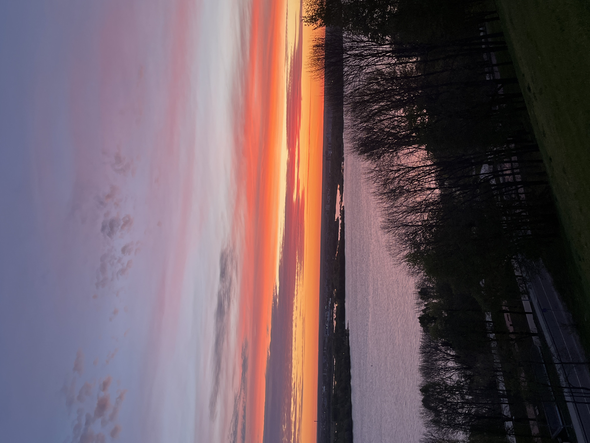 The image captures a sunset over the Volga River in Nizhny Novgorod, Russia. The sky displays vibrant hues of orange, pink, and purple. The river reflects the colors of the sunset. The silhouette of trees lines the riverbank. A road runs along the river, with occasional trees and structures visible.