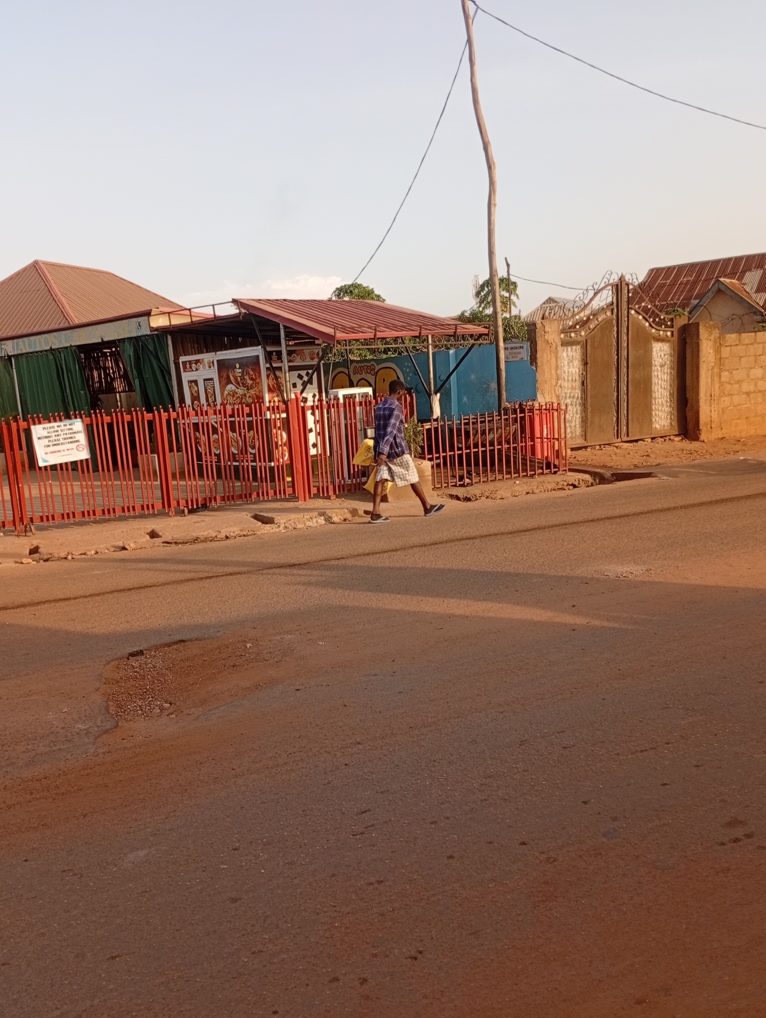 The image depicts a street scene in Narayi, Nigeria. A person wearing a blue plaid shirt and checkered shorts walks across the road carrying a yellow bag. The road is paved but has a pothole in the foreground. On the left side of the road is a building with a red fence in front of it. The building appears to be a restaurant with signs displayed. Behind the restaurant, there's a wooden post and a blue painted wall. A gate is visible further down the street, constructed of metal and concrete. The sky is light.