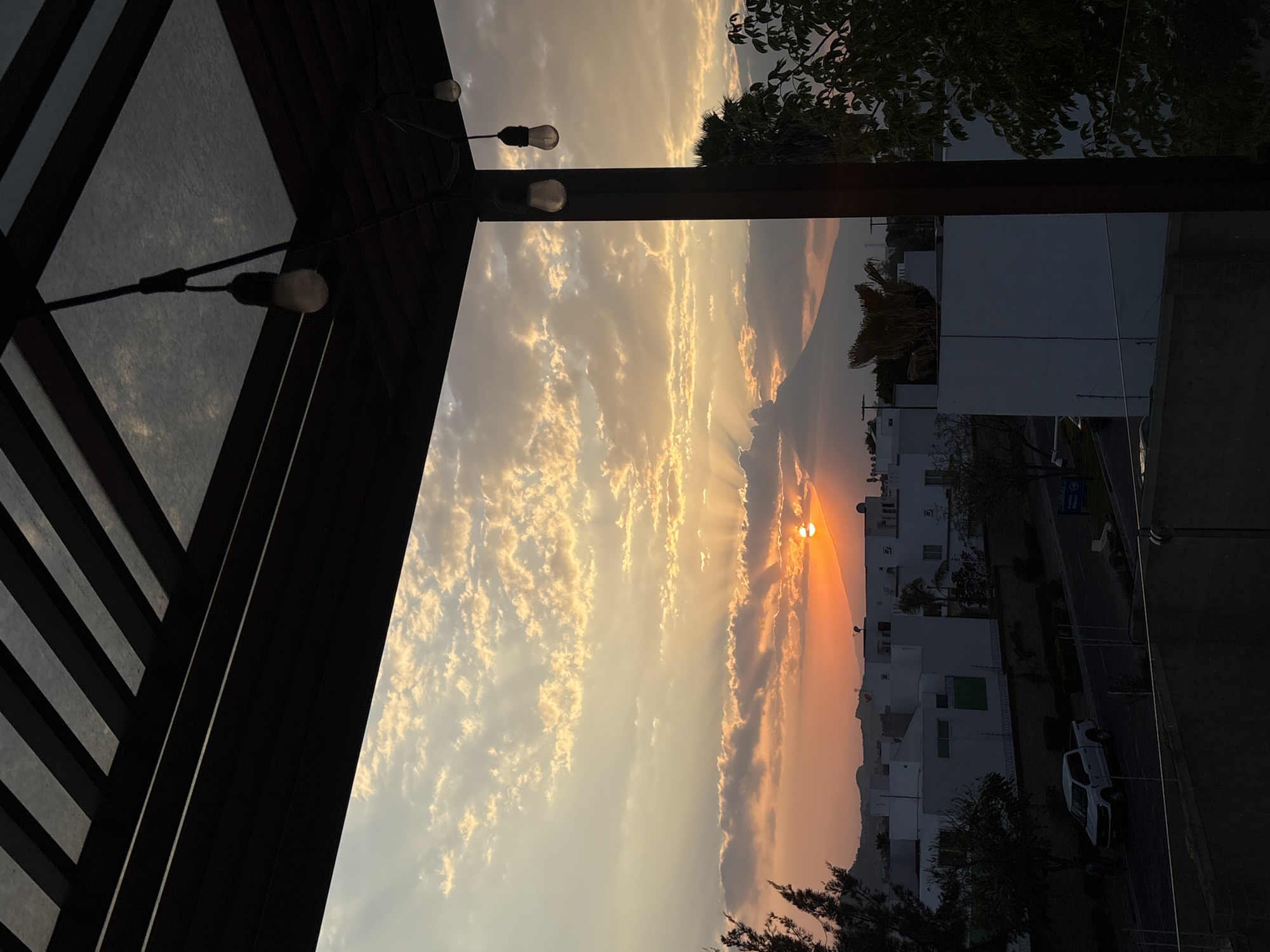 The image displays an outdoor scene at sunset in San Bernardino Tlaxcalancingo, Mexico. The perspective is from beneath a dark-colored pergola structure featuring a slatted roof with translucent panels. Multiple string lights with exposed incandescent bulbs hang from the pergola, and a dark vertical support beam is visible on the right side of the frame. In the midground, a residential area is visible, comprising several white, flat-roofed buildings and interspersed trees and shrubs. A paved street runs horizontally through this area, on which a white pickup truck is present. The background is dominated by a large, dark mountain, likely a volcano, silhouetted against the sky. The sun, a bright orange orb, is positioned above the mountain, casting crepuscular rays through scattered clouds. The clouds are illuminated in hues of orange, yellow, and grey, while the upper sky retains a lighter, less saturated tone. No people are visible.
