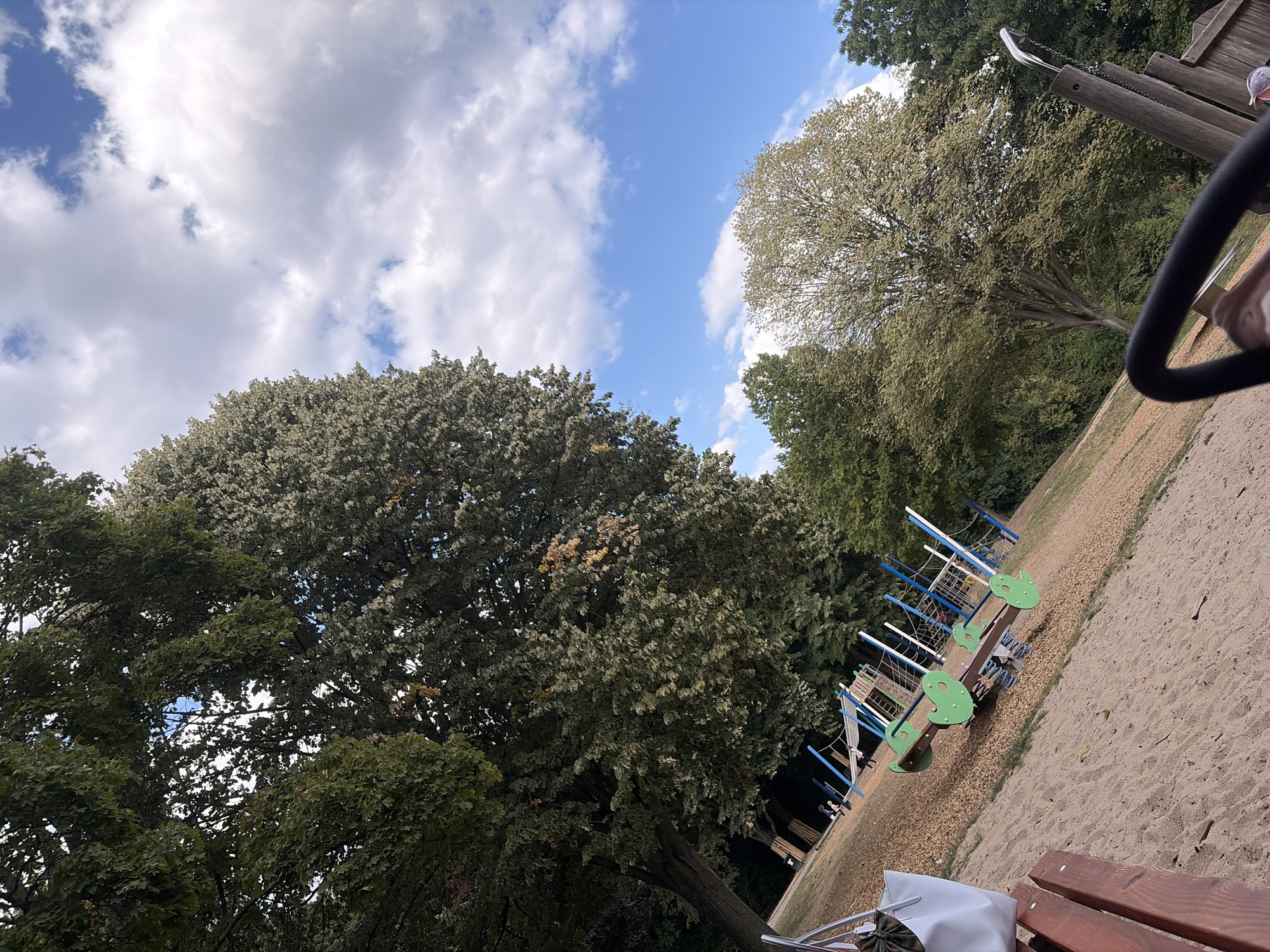 The image depicts a children's playground with a sand surface and wood chip cushioning, situated in a park setting with mature trees. The playground features a seesaw with green seats and a blue and white climbing structure. In the foreground, a portion of a park bench and a white tote bag are visible. The sky is blue with scattered white clouds. The scene is likely located in Duisburg, Germany, given the provided context.