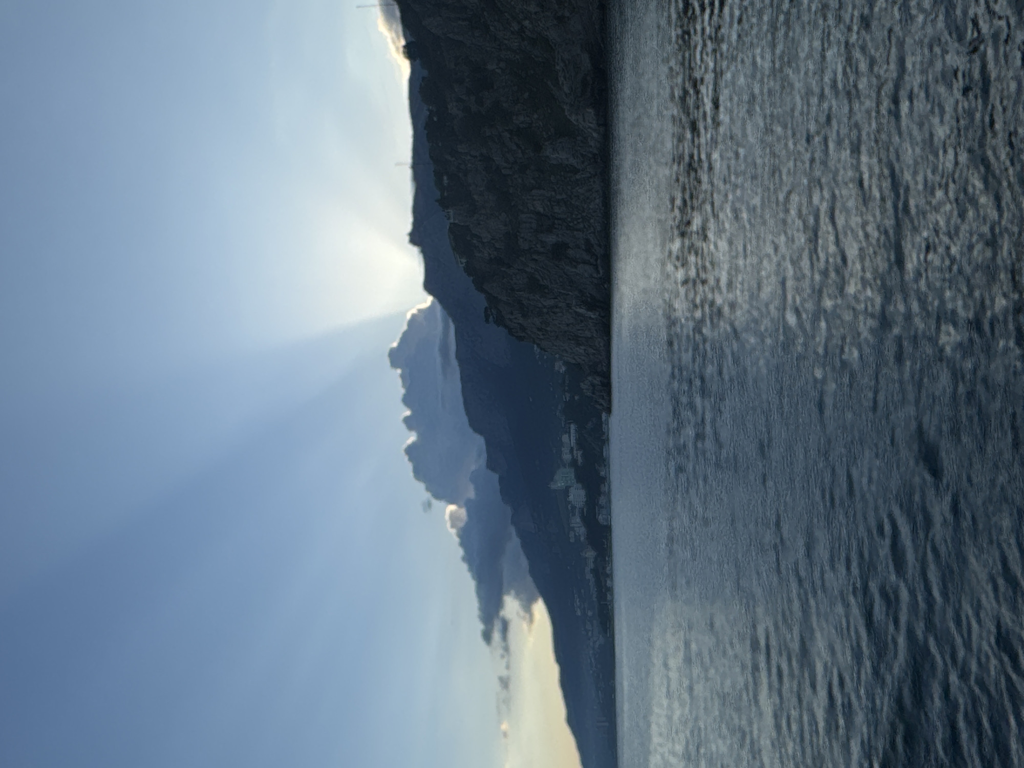 The image depicts a seascape with a rocky coastline. The sky is bright blue with visible sun rays emanating from behind a mountain or large rock formation. A body of water, likely the sea, occupies a significant portion of the right side of the image. The coastline is rugged, with dark, textured rock formations. There is evidence of habitation along the coastline, possibly buildings. The scene is located in or near Гаспринский поселковый совет, Ukraine, based on the prompt.