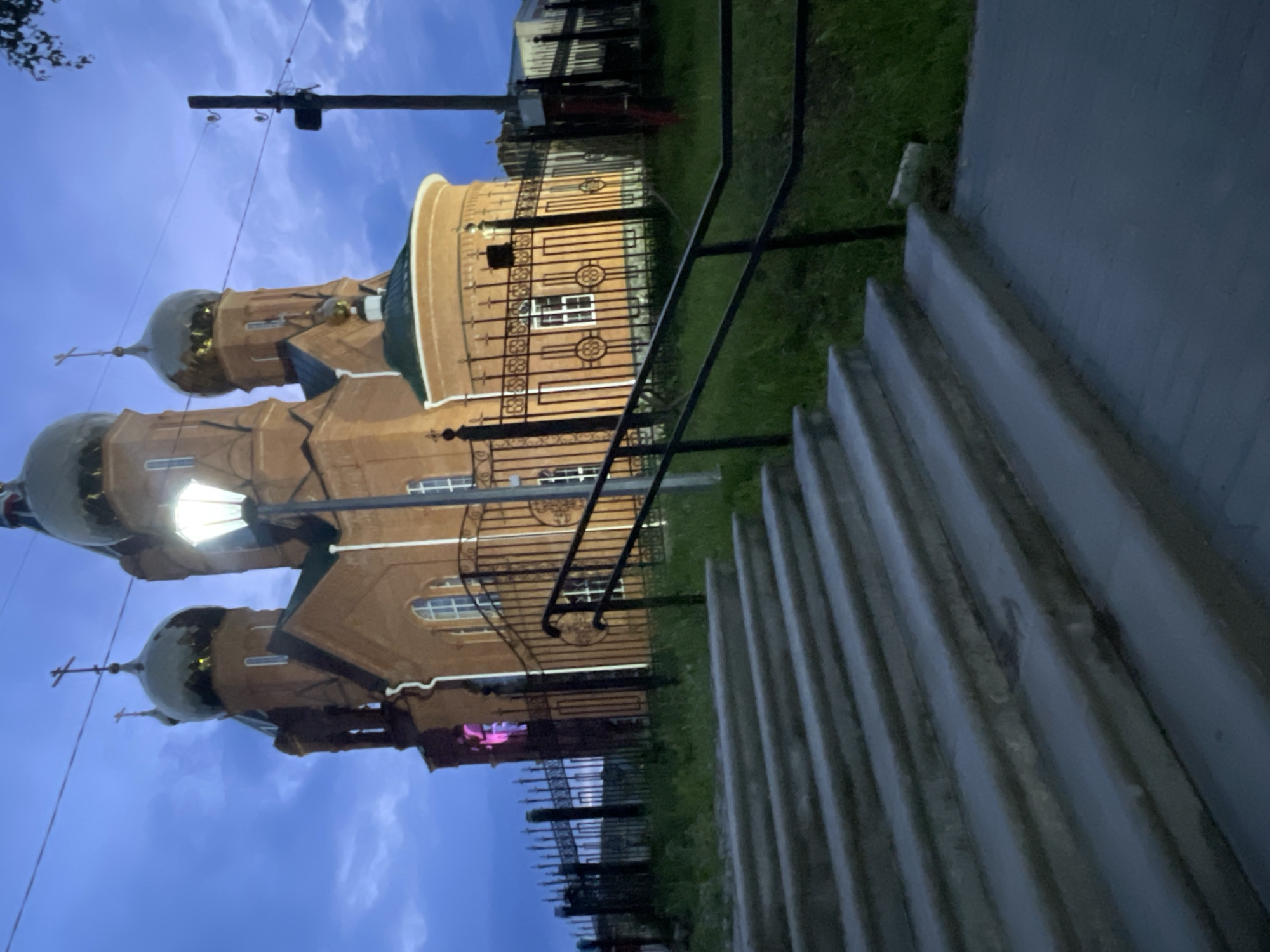 The image displays an illuminated multi-domed Orthodox Christian church at dusk or early night in Красные Баки, Russia. The church is constructed of light brown brick, featuring several greyish-green onion domes, some adorned with golden accents and crosses. One of the central domes is brightly lit, and light also emanates from various arched and rectangular windows. A vibrant pink or purple light source is visible behind a decorative black wrought-iron fence towards the lower part of the church structure.
In the foreground, a flight of concrete or stone steps ascends along the right side of the frame, bordered by a black metal handrail. This railing extends to form a fence separating the steps and a patch of green grass from the church property. A utility pole with overhead wires is visible against the deep blue, slightly clouded sky on the left. The scene is static, with no people or discernible text. The overall atmosphere is serene, highlighting the church's architecture under artificial illumination.