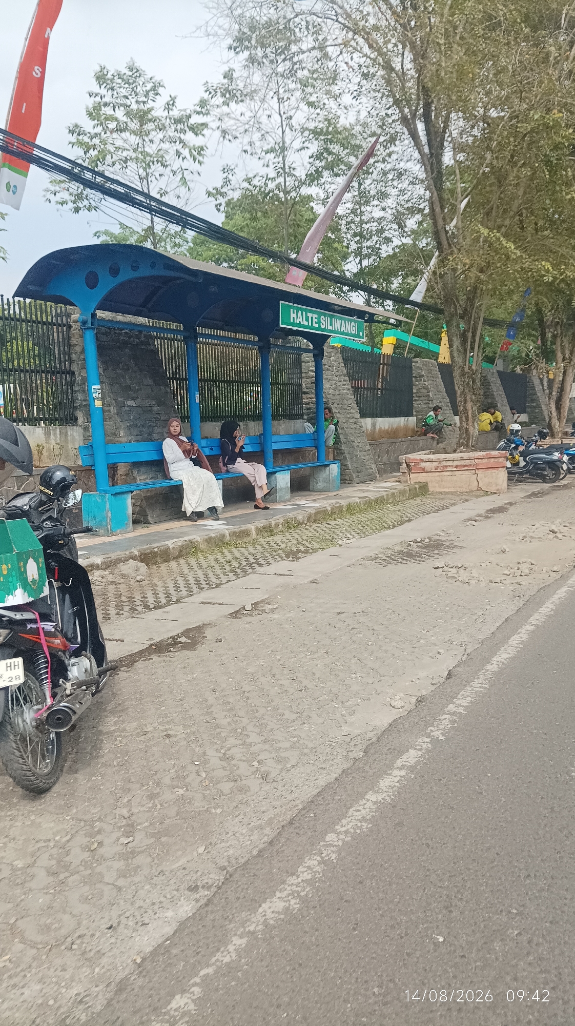 The image shows a bus stop labeled "HALTE SILIWANGI" in Tasikmalaya, Indonesia. Several people are seated on the blue benches under the blue shelter. A motorcycle with a green box attached is parked on the left side of the frame. The bus stop is situated next to a paved road and a walkway made of interlocking concrete blocks. Behind the bus stop, a stone wall and trees are visible. In the background, there are more trees and a cloudy sky. The timestamp "14/08/2026 09:42" is visible in the lower right corner of the image.
