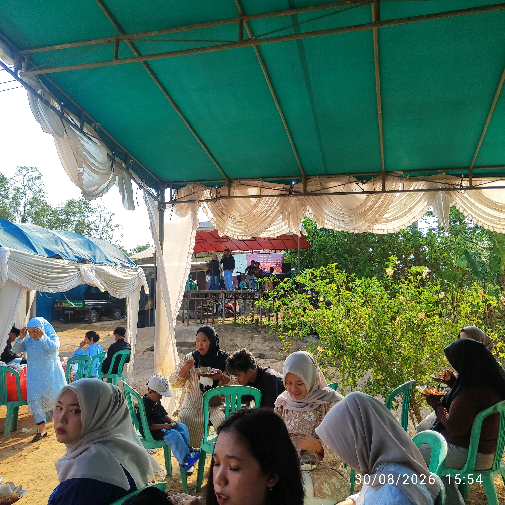 In the city of Pembaharuan, Indonesia, an outdoor gathering is taking place under a green canopy supported by a metal frame. The canopy is draped with white fabric. In the foreground, several people are seated on green plastic chairs, some eating or holding plates of food. A young woman in a light blue dress and hijab is standing and adjusting her headscarf. Behind them, a stage with a red backdrop is set up, where a band or performers are visible. A truck is parked in the background. The environment is characterized by sandy ground and lush green foliage. The timestamp "30/08/2026 15:54" is visible in the bottom right corner of the image.