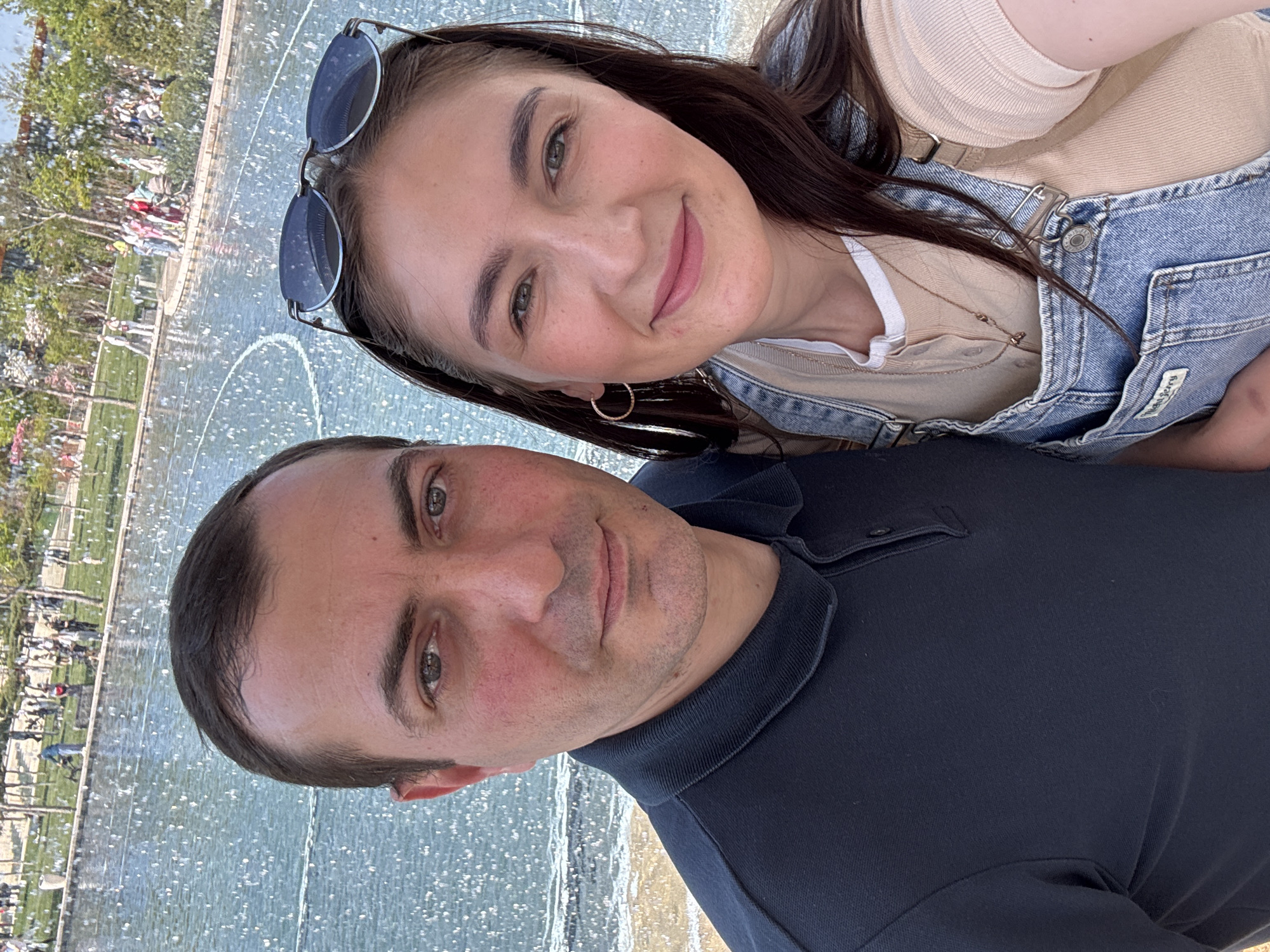 A medium shot captures two individuals posing for a selfie in front of a water fountain. The woman is positioned slightly behind and to the right of the man. She has sunglasses perched on her head, brown hair, and wears a denim jacket over a light-colored top. She is smiling and has fair skin. The man is wearing a dark blue polo shirt and also smiles at the camera.
The background is dominated by the water fountain and a park area with multiple people visible in the distance. The fountain appears to be located in a large plaza or park setting. This is located at the fountain in front of the Drama Theater in Krasnodar, Russia.