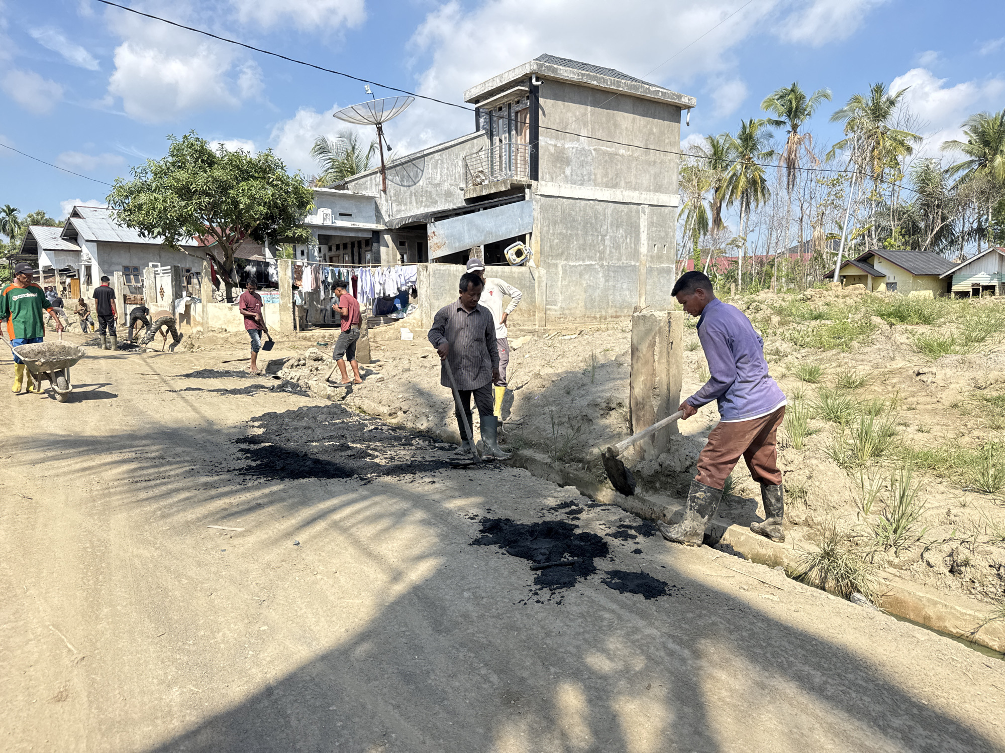 In Meunasah Raya, Indonesia, on a sunny day, several individuals are engaged in road construction work. The scene depicts a rural or suburban setting with several modest houses, some with laundry hanging outside. The foreground shows a dirt road with piles of dark material, possibly asphalt or gravel, scattered on it. Several men are actively working, using shovels and a wheelbarrow to move the material and compact the road. One man in a green shirt and yellow boots is pushing a wheelbarrow loaded with construction material. Another man in a striped shirt is using a shovel, and a third in a purple long-sleeved shirt and brown pants is also shoveling. The environment is dry and dusty, with some vegetation and palm trees visible in the background. The sky is partly cloudy. The overall impression is one of community effort towards infrastructure improvement.