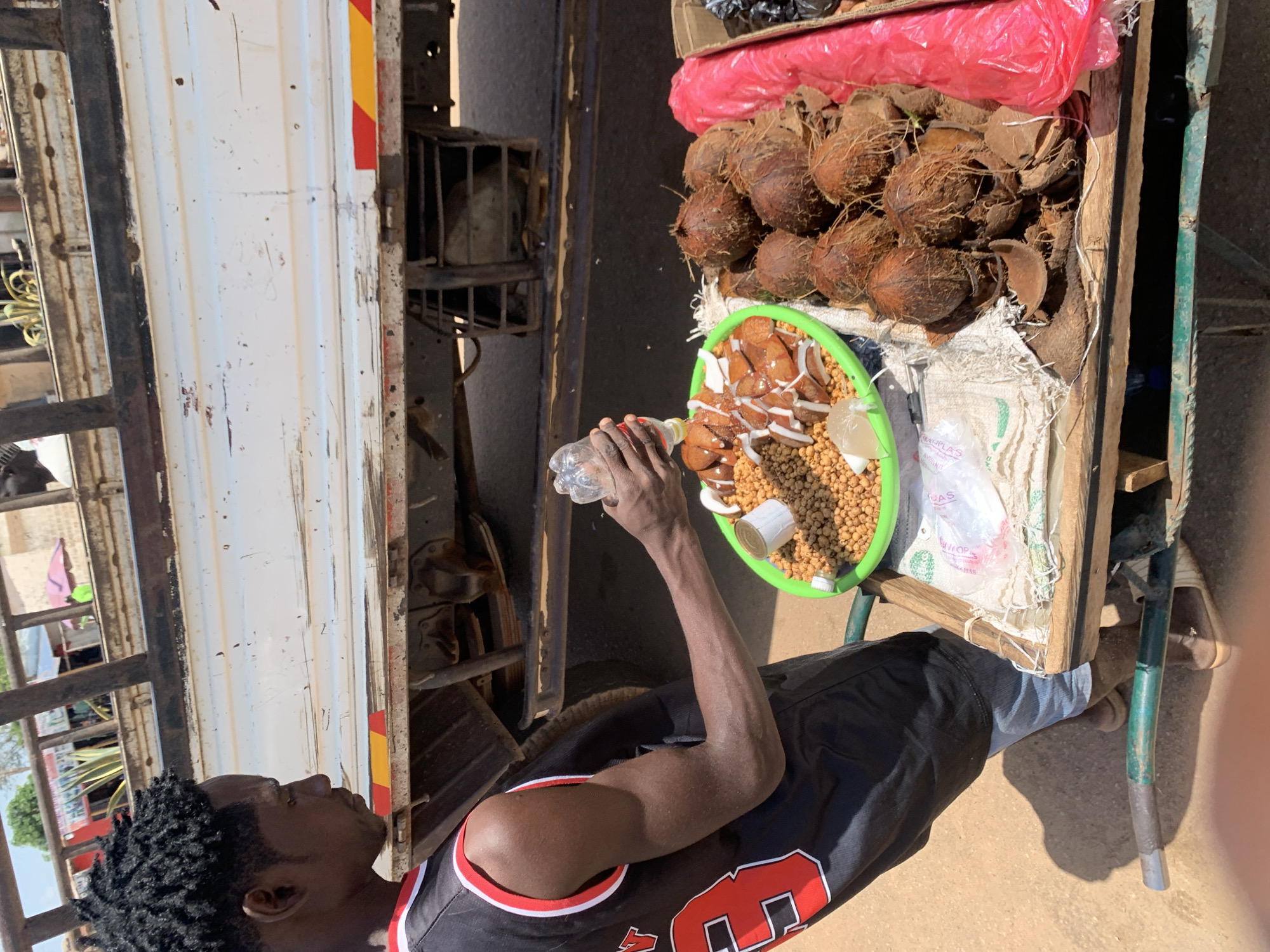 In Gura Top, Nigeria, an outdoor street or market scene unfolds under bright, sunny daylight. The primary subject is a dark-skinned man with short dreadlocks, seen from the chest up in profile. He wears a black sleeveless shirt with red and white trim on the collar and armholes, and dark pants. He stands next to a wooden vendor's cart on wheels, holding a clear plastic water bottle over a prominent green plastic tray on the cart.
The green tray contains various food items: numerous white pieces of coconut flesh, small light brown spherical grains (possibly groundnuts or millet balls), and a small clear plastic sachet. A small tin can is also visible within the tray. The vendor's cart is heavily laden with whole, husked coconuts piled up next to the tray.
In the background, to the left, a large white truck with red and yellow reflective markings is parked or passing by. Behind the cart, a dark metal cage is visible. The ground appears to be dusty, unpaved earth. Faint text "PERUPLAS" and "WHOP" are visible on a white plastic bag within the tray on the cart. The man appears focused on the items in the tray, suggesting he is either preparing the food for sale or consuming it.