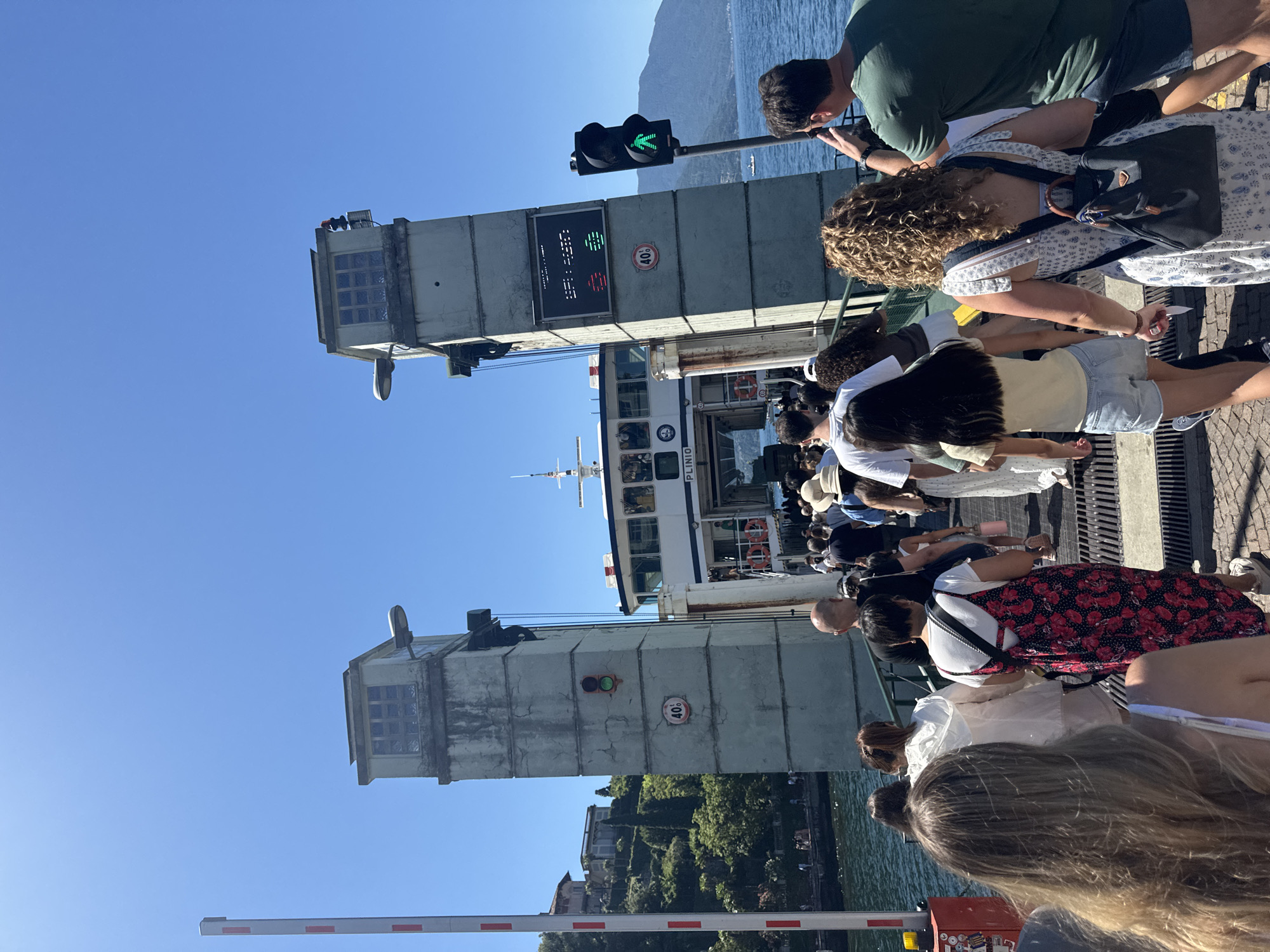 The image depicts a scene at a ferry terminal in Varenna, Italy, on a sunny day. A group of people are gathered on a walkway, waiting to board or disembark a ferry. The ferry, named "PLINIO," is visible docked at the terminal. In the background, a body of water, likely Lake Como, stretches out towards a mountain range covered in lush greenery.
Two tall concrete structures, possibly part of the ferry terminal's infrastructure or a bridge, dominate the left side of the frame. One of these structures has a traffic light with a green pedestrian signal illuminated. Below the traffic light, a digital display shows a series of symbols. A circular sign with "40%" is also attached to the structure. A white and red striped barrier is positioned at the bottom of the image, suggesting a controlled entry or exit point.
The people in the image are dressed in casual summer clothing. Their expressions are not clearly discernible, but the overall atmosphere appears to be one of anticipation or leisure, typical of a tourist destination. The sunlight suggests it is likely daytime, and the clear blue sky indicates fair weather. The presence of the ferry, the lake, and the mountainous landscape firmly places the scene in a scenic, lakeside Italian town.