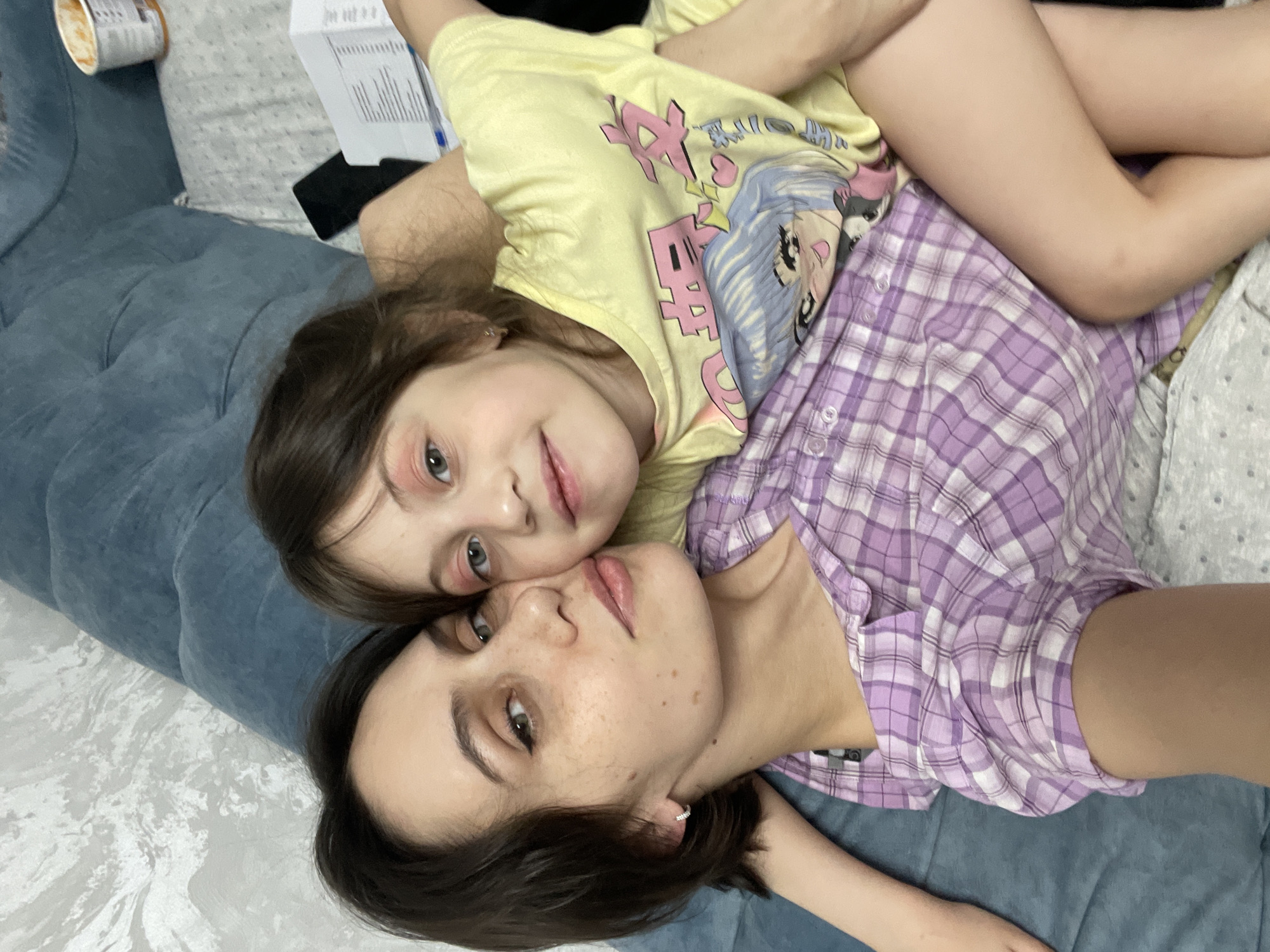 This image, taken in Izhevsk, Russia, depicts an adult female and a young female child in a close, tender moment, lying down together in an indoor setting. The photograph is oriented diagonally, showing the subjects from a slightly elevated perspective.
The child, with light brown hair and blue eyes, is resting on top of the adult. She wears a yellow t-shirt adorned with an anime-style character and stylized Japanese text. Her expression is calm, with a slight, gentle smile, and her arms are around the adult's upper body.
Beneath her, the adult female, with dark brown hair and dark eyes, wears a purple and white plaid button-up shirt. Her face shows a neutral, content expression, and she has visible freckles. A small silver hoop earring is visible in her left ear. Her bare legs are partially visible, crossed, and extend towards the upper right of the frame.
The setting appears to be a casual home environment, possibly on a couch or bed, indicated by the blue upholstered furniture on the left and a light-colored patterned sheet on the right. In the background, a cup (possibly instant noodles) and a white document with unreadable text are visible, suggesting everyday activities. Both individuals appear relaxed and comfortable, conveying a strong sense of affection and bonding. No specific time of day or weather can be discerned from the indoor scene.