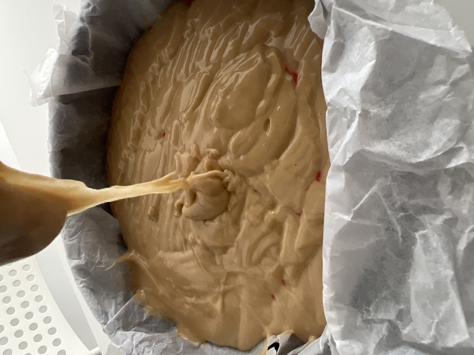 A close-up shot shows batter being poured into a round baking pan lined with parchment paper. The batter is light brown with some red specks and has a thick, swirled texture. The parchment paper is wrinkled and extends beyond the rim of the pan. A small portion of a white plastic surface with circular cutouts is visible in the lower-left corner. The setting appears to be indoors. There is no indication of the location being Mogán, Spain.