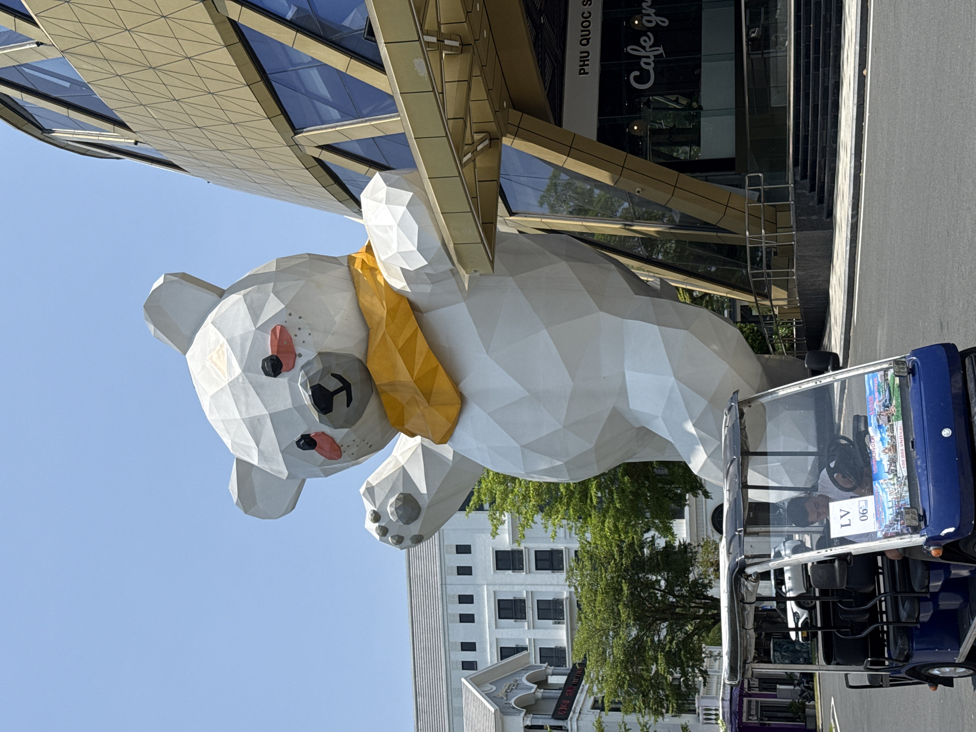 The image captures a bright outdoor scene, likely in a public or commercial area in Phú Quốc, Vietnam, under a clear blue sky. The main subject is a colossal, white, low-polygonal style bear statue, featuring a yellow scarf, black eyes and nose, and red cheeks. The bear is depicted in a dynamic, outstretched posture, suggesting a playful or welcoming gesture.
Behind the bear statue stands a modern, architecturally distinctive building characterized by geometric golden-toned panels and large reflective glass windows. Text visible on this building includes "PHU QUOC S" and "Cafe gr," likely part of "Cafe grande" or similar. To the right of the statue, a blue electric golf cart is parked, with a person visible in the driver's seat. The golf cart displays text such as "LV 06," "PHU QUOC," and pricing information "100.000 VND/car," along with fragmented words like "TOURIE," "NDER," and "SA," indicating a tourist transport service.
In the background, a multi-story white building with numerous dark windows is partially visible, interspersed with green trees. The scene is bathed in bright daylight, suggesting good weather. The unique art style of the bear statue and the surrounding elements create a vibrant, tourist-friendly atmosphere.