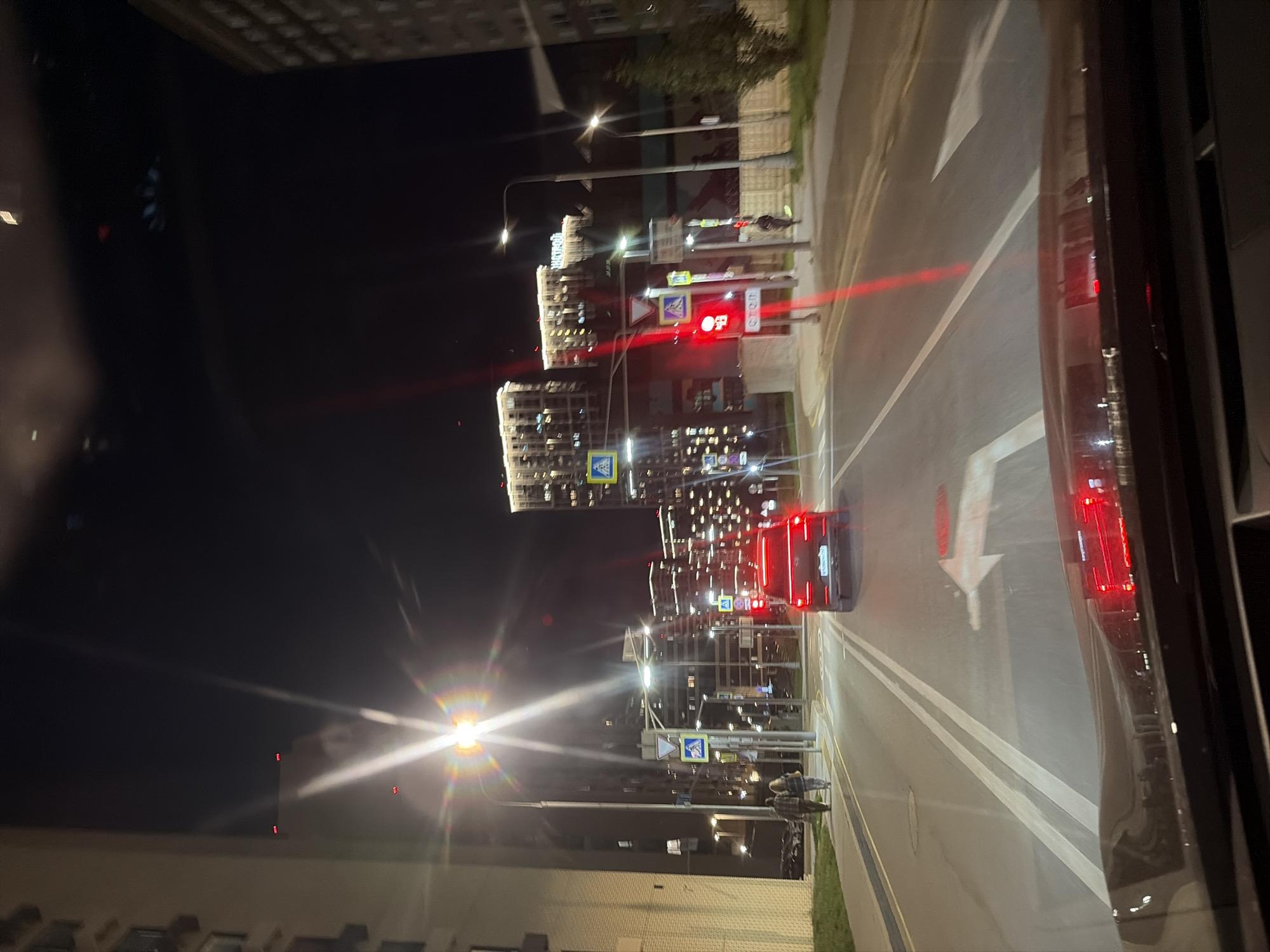 The image captures a nighttime street view in Moscow, Russia, from the perspective of someone inside a vehicle. The scene is illuminated by streetlights and the lights of residential buildings. In the foreground, the dashboard of the vehicle is visible, reflecting red lights. The road ahead shows lane markings and a directional arrow. A red car is driving in the same direction, with its taillights illuminated. Two pedestrians are walking on the sidewalk to the left of the road. Several tall residential buildings with many windows lit up line the street. Traffic signals and street lamps are also present. The overall atmosphere is urban and nocturnal.