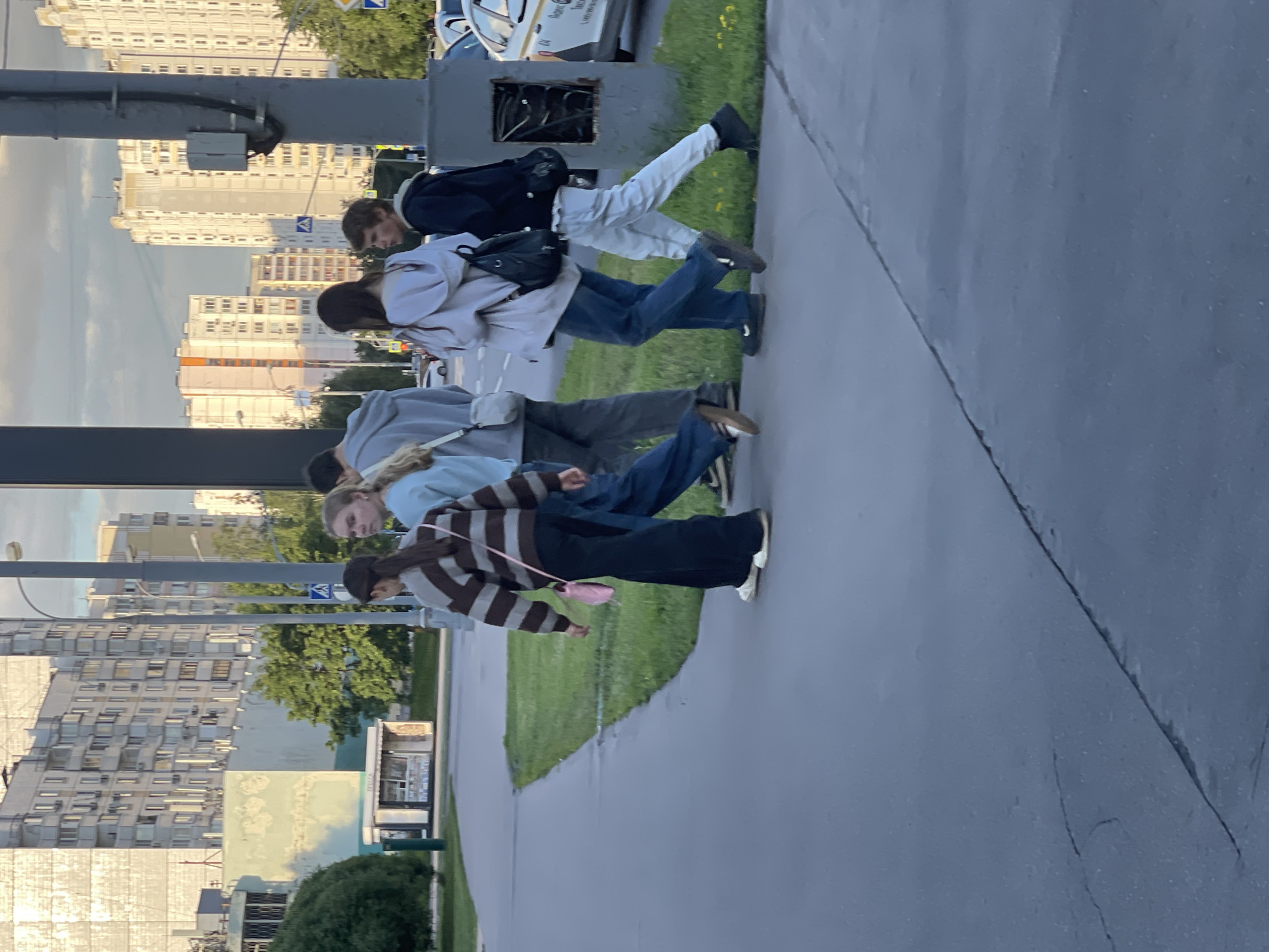 A group of young people are walking along a paved path beside a grassy area. The scene is urban, with tall, modern apartment buildings visible in the background. The sky is overcast, suggesting a cloudy day. The people are dressed casually in jeans, hoodies, and jackets. Some are carrying backpacks. The overall impression is of people going about their day in a city environment. There are no clear indications of a specific event or strong emotions displayed. The time of day appears to be daytime, possibly late afternoon, given the lighting. Visible text on a distant vehicle reads "04". The location is identified as Moscow, Russia, which is consistent with the architecture of the buildings.