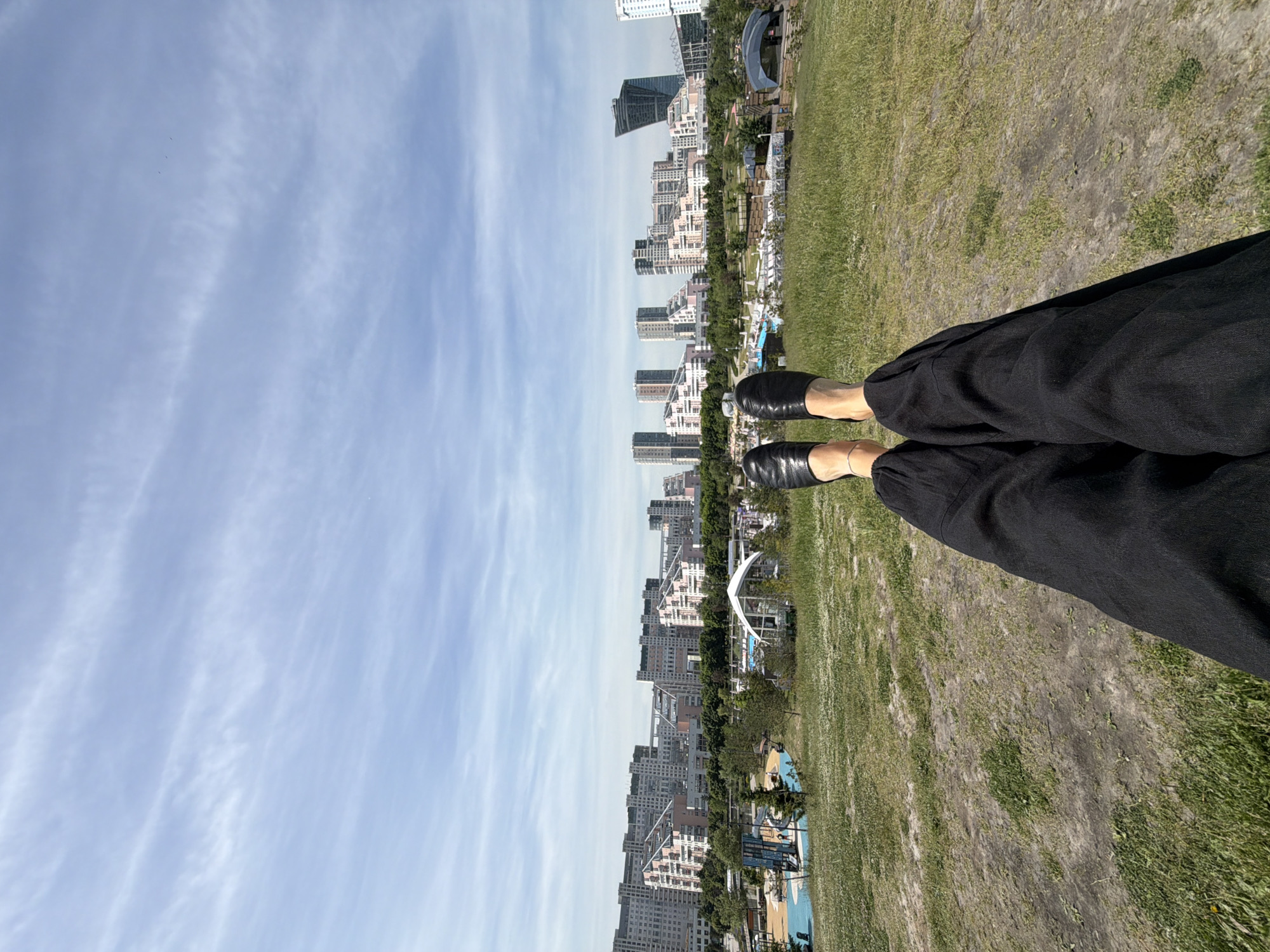 The image captures a first-person, eye-level perspective from an individual resting on a grassy incline, looking out over a park toward a cityscape identified as Khimki, Russia.
In the immediate foreground, the viewer's legs are extended horizontally, dressed in black wide-leg trousers and black slip-on shoes. A slender silver ankle bracelet adorns the right ankle. The ground directly beneath the viewer is a patchy mix of green grass and areas of dry, exposed soil.
The midground consists of a verdant grassy slope, punctuated by scattered white wildflowers and some drier patches. Beyond this slope, a recreational park area is visible, featuring a brightly colored blue playground, numerous trees and shrubs, and several white, tent-like structures, including one with a distinct arched roof.
The background presents the urban skyline of Khimki, Russia. A prominent residential complex with distinctive multi-story buildings exhibits a stepped or terraced architectural design, characterized by light-colored facades with sections of soft pink or salmon. Interspersed are taller, modern high-rise structures, some with reflective glass exteriors. On the far right, a dark, angular skyscraper with a unique, geometric design stands as a landmark, identifiable as the Khimki Business Centre. Roads and infrastructure are visible between the buildings.
The sky above is a clear light blue, streaked with thin, horizontal white cirrus clouds. Three small, dark bird-like figures are discernible in the upper right quadrant of the sky. The scene is brightly illuminated by daylight.