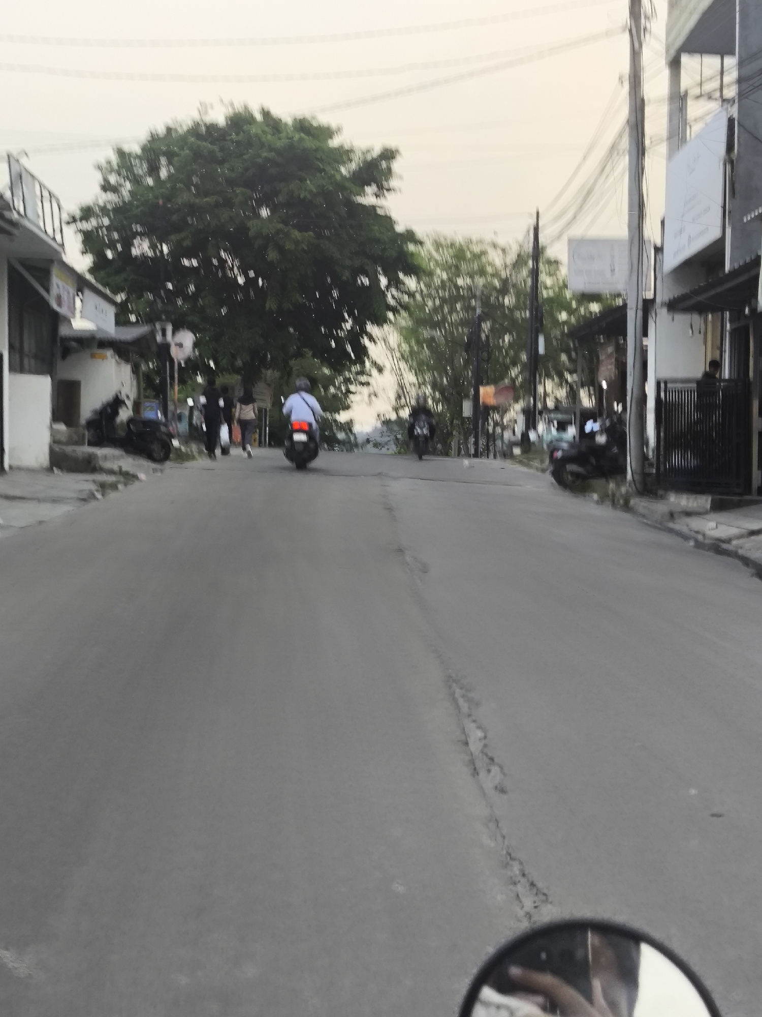 The image depicts a street scene in Cilegon, Indonesia. The perspective is from a moving vehicle, likely a motorcycle, as evidenced by the presence of a rearview mirror in the foreground. The road is paved and appears to be in fair condition, with some visible cracks and unevenness. Several individuals are present, some walking and others on motorcycles. On the left side of the road, there are buildings with storefronts, and a large tree provides shade. On the right side, more buildings and a black metal fence are visible. The sky is overcast, suggesting it could be early morning or late afternoon. The overall atmosphere is that of an ordinary urban street in Indonesia.