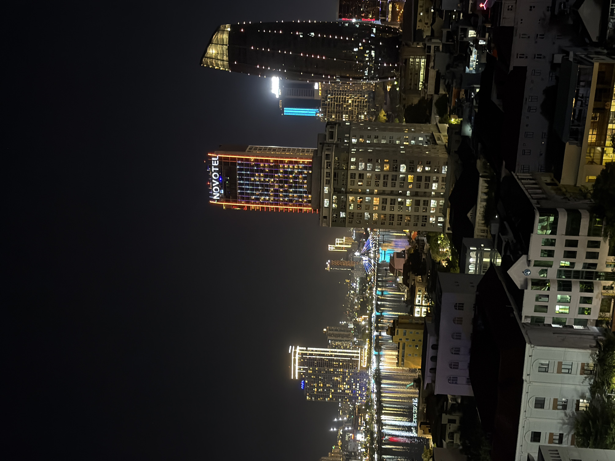 The image shows a nighttime view of the city of Đà Nẵng, Vietnam, with a focus on its waterfront skyline. In the foreground, several buildings are visible, some with their windows illuminated. In the middle ground, a river or bay reflects the city lights, creating a shimmering effect. Dominating the skyline are several tall buildings, including the Novotel Danang Premier Han River hotel, identifiable by its illuminated "NOVOTEL" sign. Another prominent skyscraper to its right has a dark, curved facade studded with lights. Across the water, other illuminated buildings and structures, possibly bridges, contribute to the vibrant cityscape. The overall impression is one of a modern, bustling city at night, with its architecture and lights creating a dynamic visual spectacle.