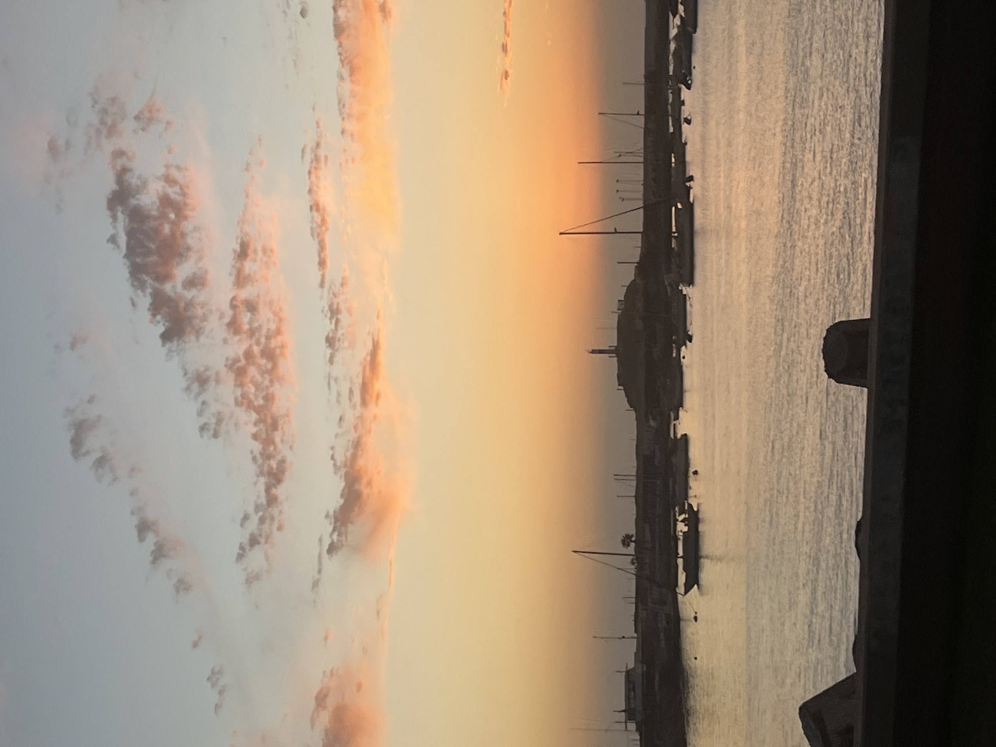 The image shows a harbor scene at sunset in Arica, Chile. The sky is a mix of light blue and orange hues with pink-tinted clouds. The sea reflects the colors of the sky. Silhouettes of boats and buildings line the harbor's edge. A dark structure is visible on the right edge of the frame, possibly part of a building or a balcony.