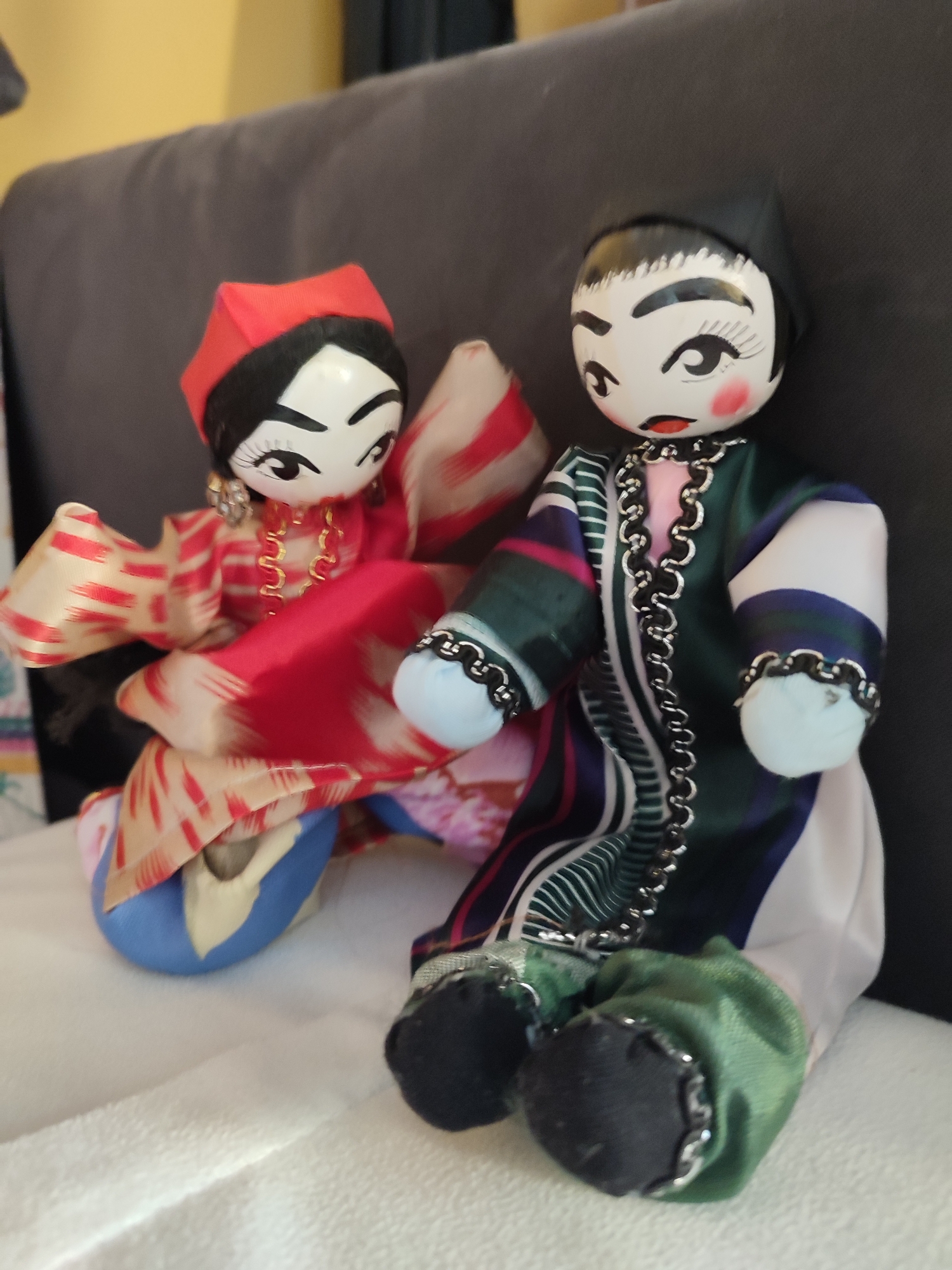 The image shows two dolls with painted faces and traditional clothing. The doll on the left is wearing a red head covering and a red and white patterned robe. The doll on the right is wearing a black head covering and a green robe with colorful stripes and silver trim. Both dolls are sitting down, and their clothing is made of shiny fabric. The dolls are positioned on a soft, white surface, possibly a blanket or upholstery, with a dark gray surface behind them. The lighting suggests an indoor setting. The scene does not depict any specific location in Üsküdar, Turkey, or any actions or interactions other than the static pose of the dolls.