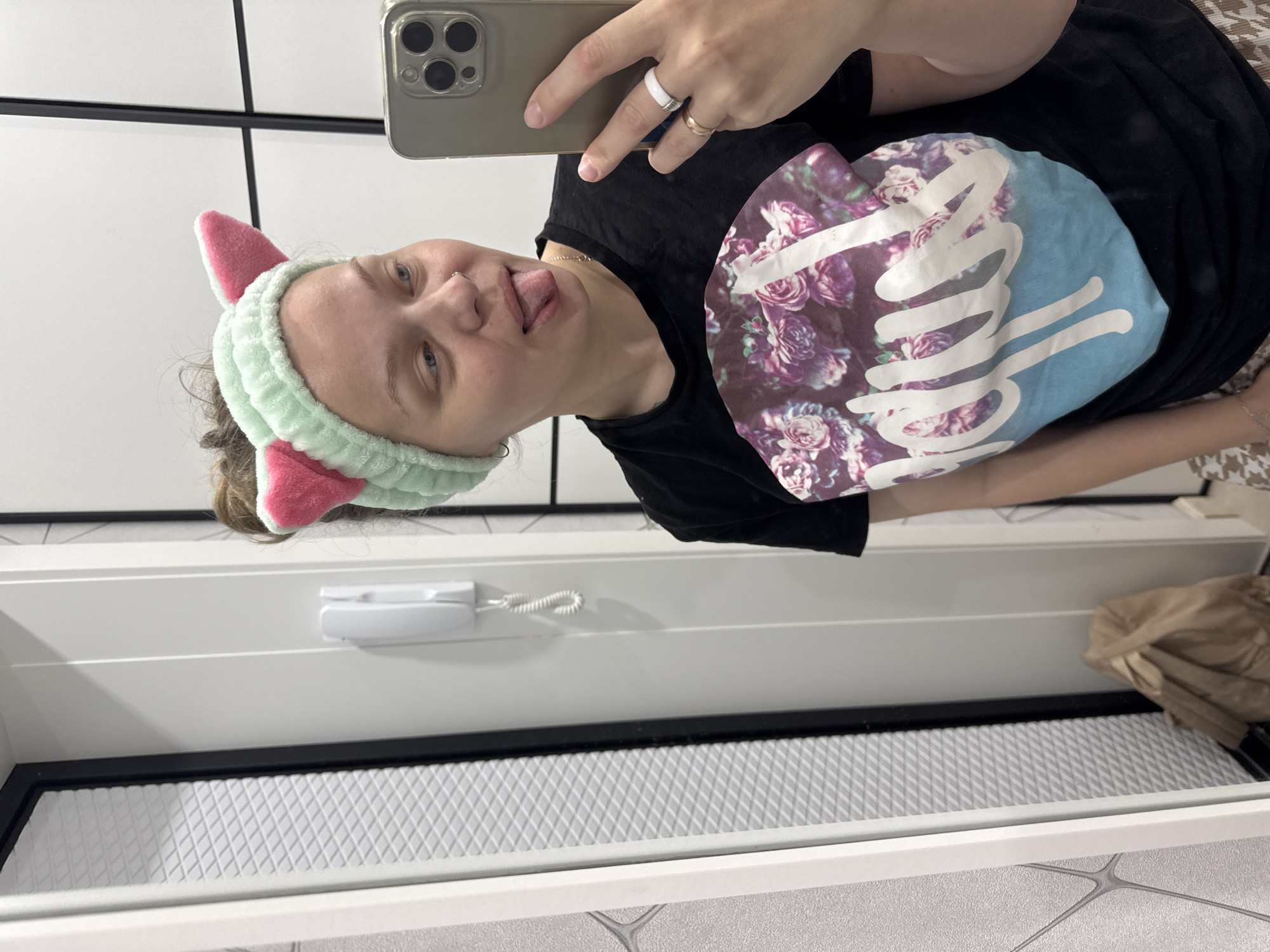A young woman is taking a selfie in what appears to be a bathroom or dressing area. She is wearing a mint green headband with pink cat ears and a black t-shirt with a floral graphic. Her tongue is sticking out. She is wearing two rings on her right hand, which is holding a smartphone. The background shows a white wall with black trim, a white intercom system, and a beige bag on the floor. The location is identified as Mikhaylovsk, Russia.
