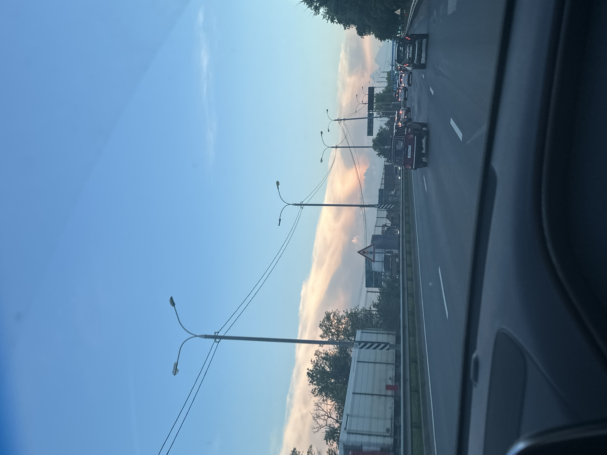 An outdoor, eye-level, vertical, medium shot captures a view of a highway from the perspective of a vehicle's interior dashboard. The highway is busy with traffic, including a red car in the foreground and several other vehicles further down the road. Overhead, streetlights and power lines crisscross the sky. In the distance, the sky is filled with soft, pale pink and orange clouds, hinting at sunset. The environment appears to be Aksay, Russia, based on the visible signs and license plates, suggesting a suburban or semi-rural highway setting. The lighting suggests it is late afternoon or early evening.
