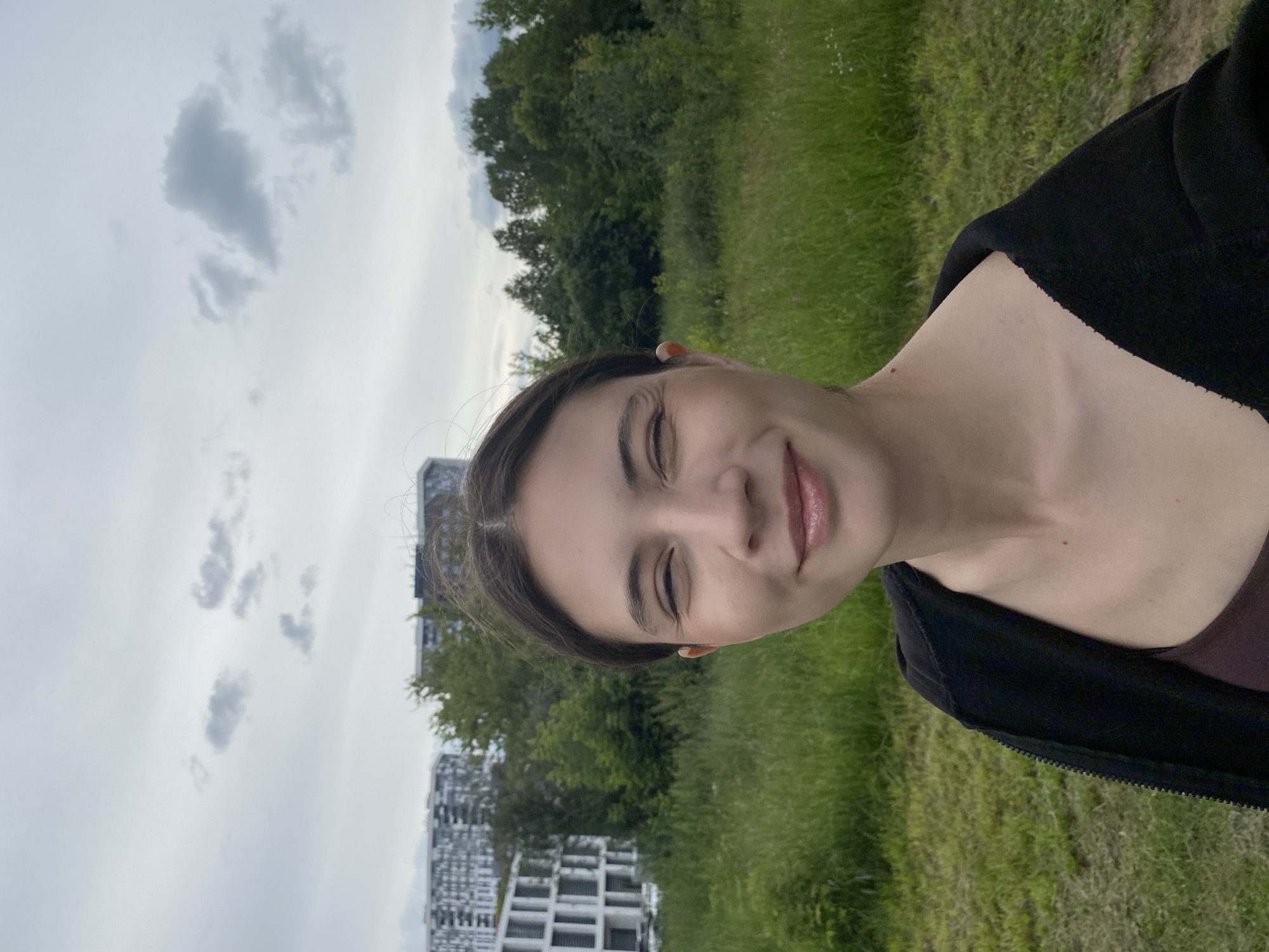 A young woman with dark hair, wearing a dark-colored zip-up hoodie over a dark tank top, stands in an outdoor grassy area. She is smiling with her eyes squinted and has lip gloss on. In the background, there are modern buildings, trees, and a cloudy sky. The location is an outdoor area in the city of Izhevsk, Russia.