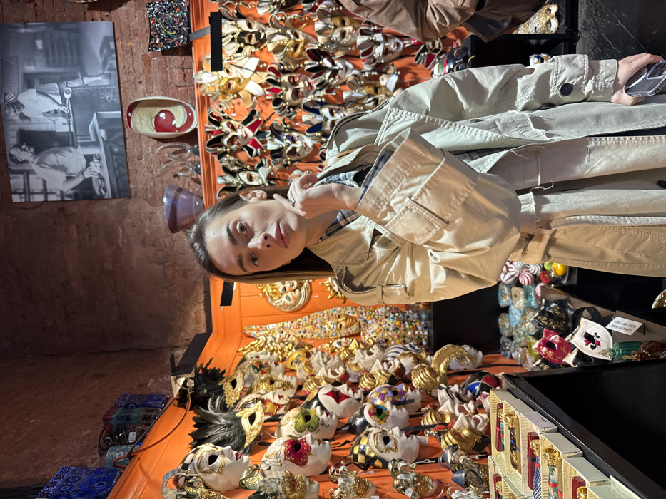 The image captures the interior of a retail shop, specifically a mask and possibly glass artisan store, located in Venice, Italy. A young adult female, with light skin and dark, straight hair, stands in the center foreground, directly facing the camera with a neutral expression. She is dressed in a light-colored trench coat worn over a plaid shirt. Her left hand is raised, with fingertips resting near her chin, while her right hand grips a dark-colored smartphone.
The background is densely populated with a diverse array of Venetian masks, characteristic of the Carnivale di Venezia, displayed on orange-painted walls and shelves. These masks vary in design, color, and embellishment, featuring gold accents, intricate patterns, and feathers. Above the mask displays, the upper wall consists of exposed reddish-brown brick. Affixed to the brick wall is a large black and white photograph depicting two individuals engaged in glassblowing, indicating a connection to Murano glass. Below this photograph, various decorative glass objects, including a dark purple bowl and a red and white oval dish, are visible on a shelf. To the left, a black display unit holds rows of boxed merchandise. A small portion of another individual wearing a brown jacket is partially visible on the far right edge of the frame.