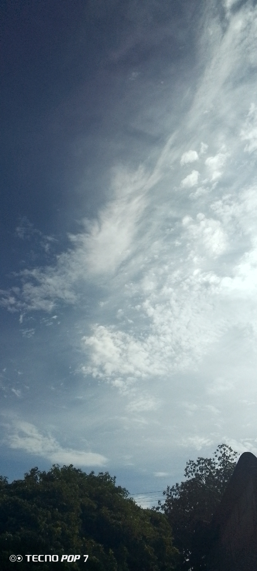 The image depicts a daytime view of the sky in Altagracia, Venezuela. The upper left portion of the sky is a deep blue color, transitioning to a lighter blue with varying cloud formations across the rest of the sky. These formations include cirrus and cumulus clouds. At the bottom edge of the frame, there's a visible treeline with dense foliage. At the lower left-hand corner is the written text "Tecno Pop 7."