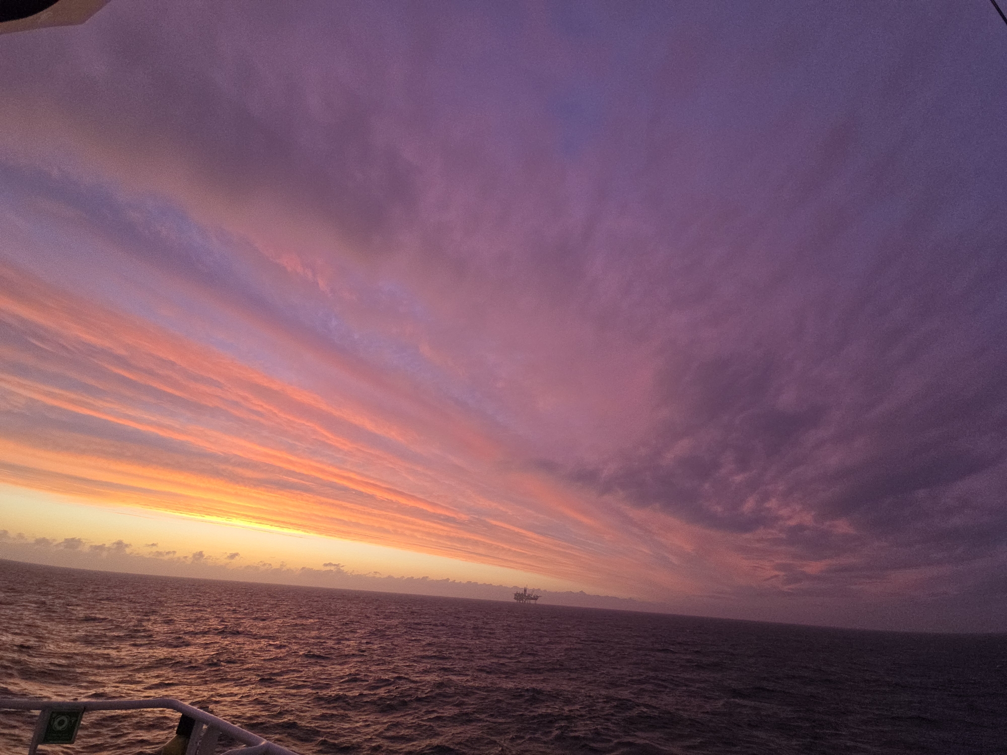 A wide-angle shot depicts a vivid sunset over the ocean. The sky is dominated by linear, horizontal clouds streaked with oranges and pinks near the horizon, transitioning into shades of purple and mauve further above. The sea mirrors these colors with gentle waves visible. In the distance, a dark silhouette, possibly an oil rig or a ship, can be seen on the horizon. The lower left corner shows a part of the ship's railing with a green safety label. The location is at sea.