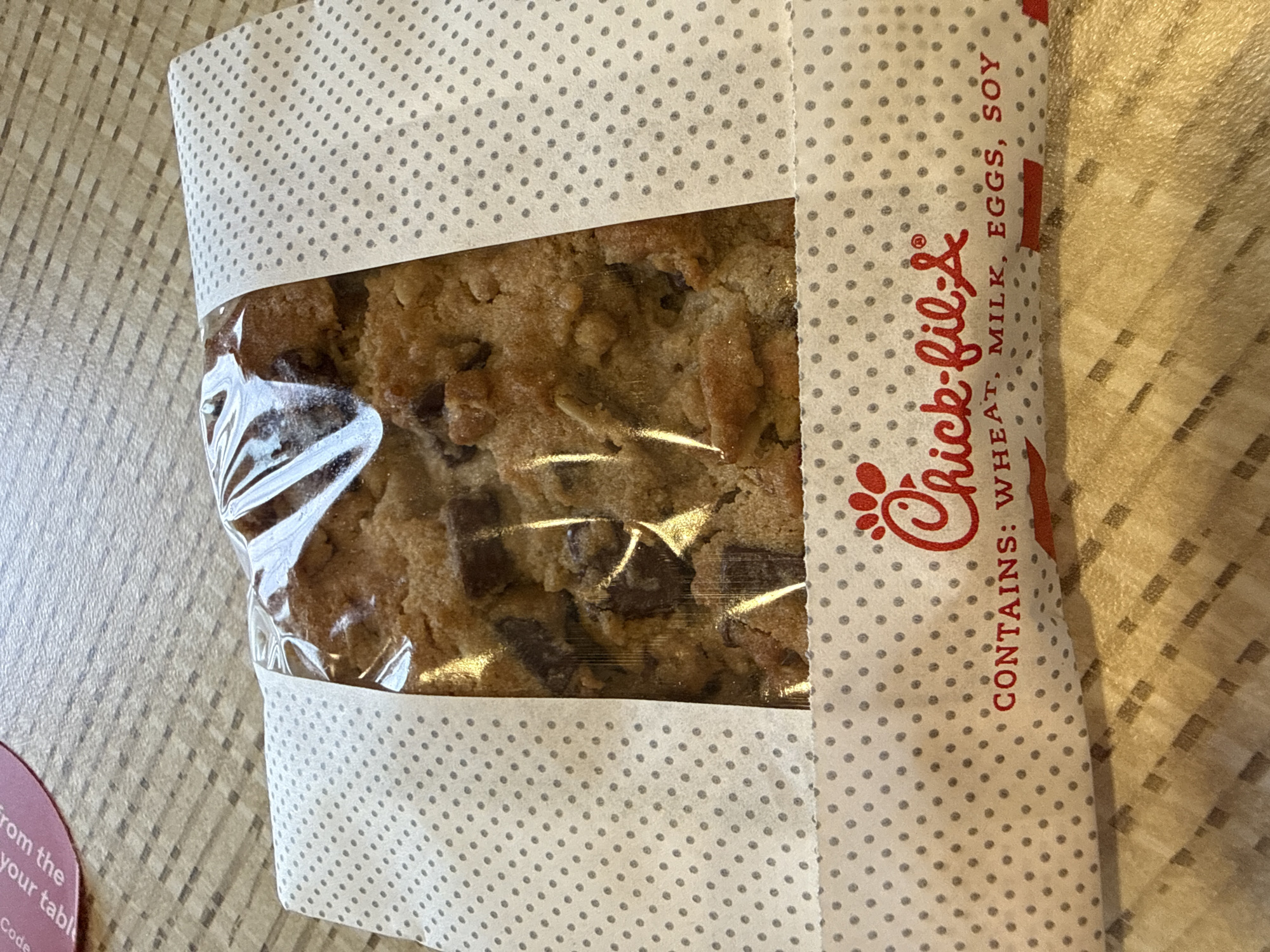 The image shows a packaged chocolate chip cookie from Chick-fil-A. The packaging is primarily white with gray polka dots, and a transparent section revealing the cookie inside. The Chick-fil-A logo is printed in red on the packaging, along with an ingredients list: "CONTAINS: WHEAT, MILK, EGGS, SOY." The cookie appears to be enclosed in clear plastic within the packaging. The package sits on a surface with a grid pattern. A small portion of a pink circular object is visible in the lower left corner, marked with the words "from the your tabl Code."