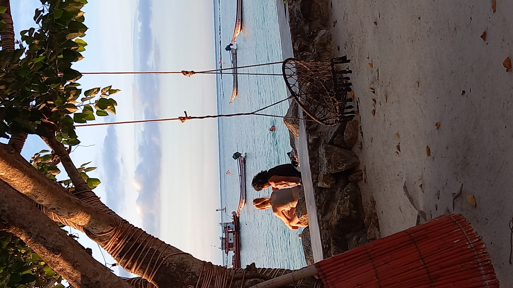 The image depicts a serene beach scene in Koh Phangan, Thailand. In the foreground, a large tree with lush green leaves is partially visible, its branches adorned with coils of rope. Two people are seated on rocks by the water's edge. One person, with dark curly hair, is wearing a black top and is looking down. The other person, wearing a light-colored top, is looking towards the sea.
The background reveals a calm, blue ocean dotted with several traditional Thai longtail boats. The water appears tranquil, and a person can be seen swimming in the distance. The sky is a soft gradient of blue and white, suggesting it might be late afternoon or early evening. The overall atmosphere is peaceful and relaxed, typical of a tropical beach setting. The ground appears to be a mixture of sand and rocks, with some fallen leaves scattered around. A woven mat, possibly a sunshade, is visible in the bottom right corner of the frame.