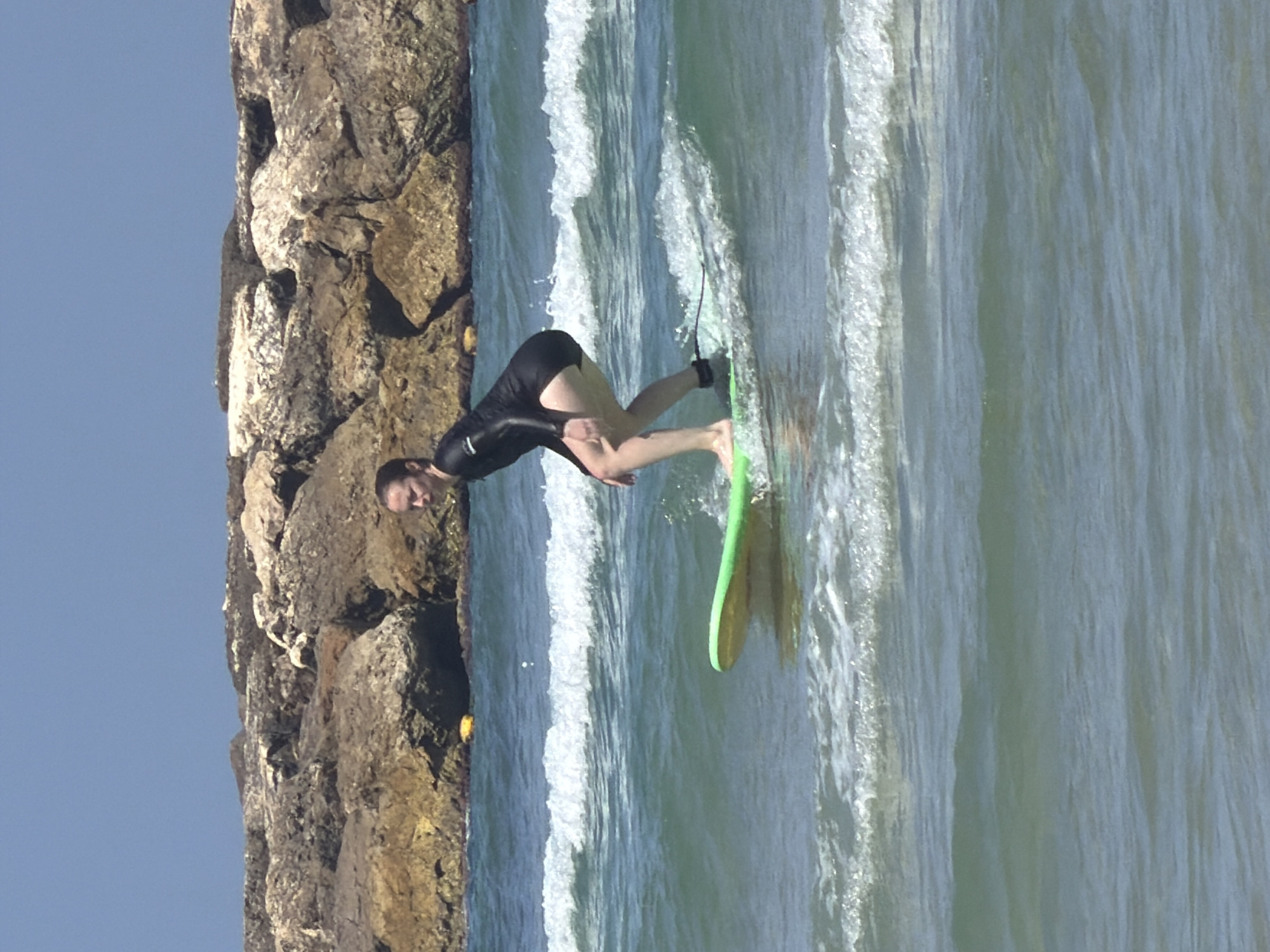 A person is surfing in the sea, near a rocky barrier. The person is wearing a black wetsuit and is riding a green surfboard. The sea has some white waves, and the sky is blue. The location is presumed to be in Tel-Aviv, Israel, based on the image's origin.