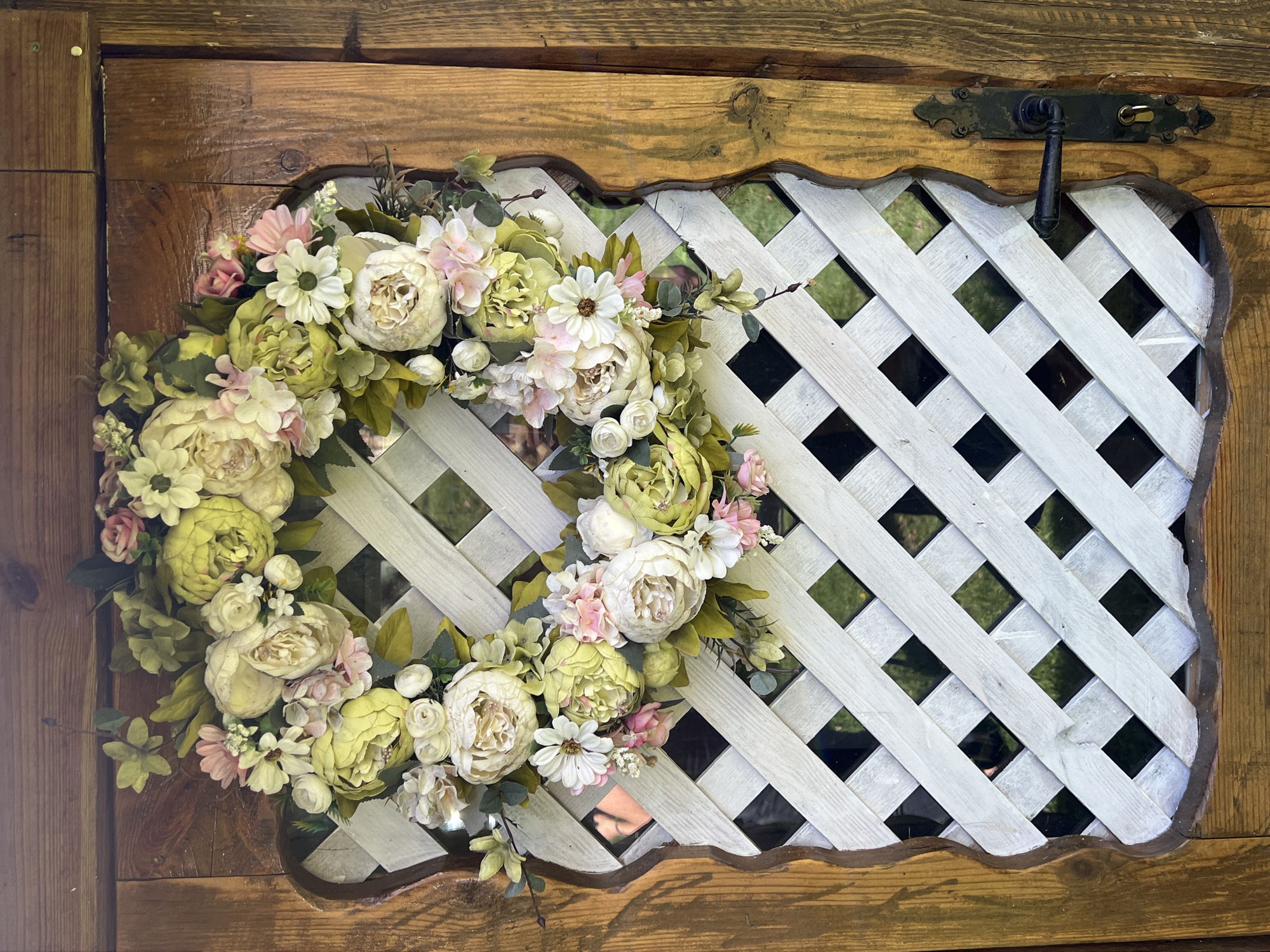A floral wreath is displayed on a weathered wooden door. The door has a lattice-like opening in the center, painted white, through which glimpses of greenery are visible. The wreath is composed of a variety of artificial flowers in shades of cream, pale green, and pink, interspersed with foliage. The door itself appears to be an exterior door, possibly of a shed or outbuilding, given its rustic appearance and the presence of a black metal door handle and lock. The scene is illuminated by natural light, suggesting an outdoor setting in Daulėnai, Lithuania.