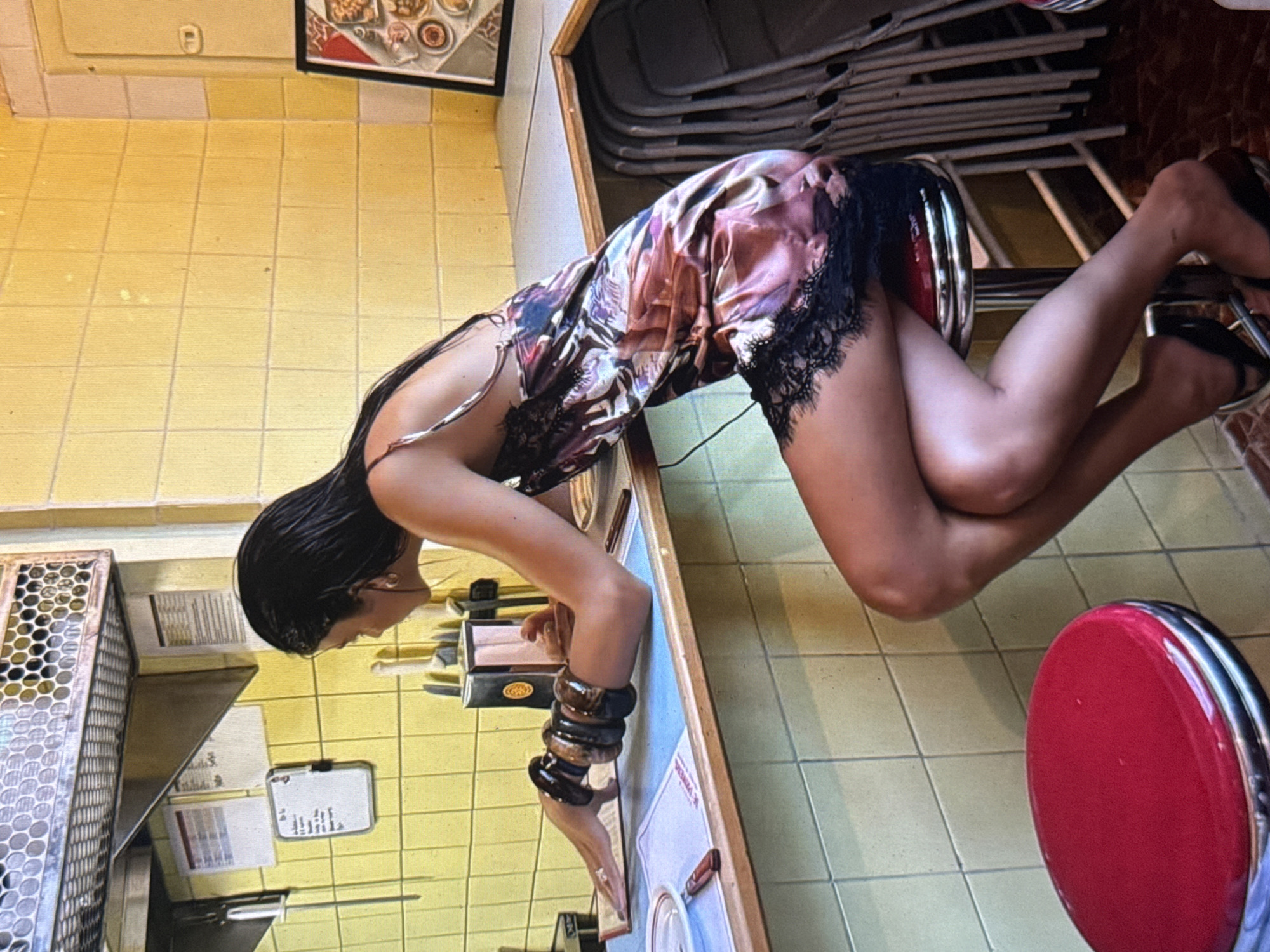 A woman with dark hair, wearing a patterned slip dress with black lace trim and dark heels, is captured in a pose where she is bent over a counter. Her left arm is resting on the counter, adorned with several large, dark, wooden bangles. The counter is reflective, showing a mirrored image of the scene, including the woman's face and the patterned tiles on the wall.
The setting appears to be indoors, possibly a cafe or diner, indicated by the tiled walls and floors, a metal rack with circular openings, and stools with red seats. There is a framed picture on the wall in the background. The lighting suggests it could be daytime. The woman's posture and the overall ambiance do not convey a strong sense of emotion, appearing more casual and perhaps posed. No visible text is clearly legible in the image, although there appear to be some labels or notes on the counter and the wall. The provided location context is Kuta Selatan, Indonesia, which might inform the architectural style or decor, though specific Indonesian cues are not overtly apparent from the visual information alone.