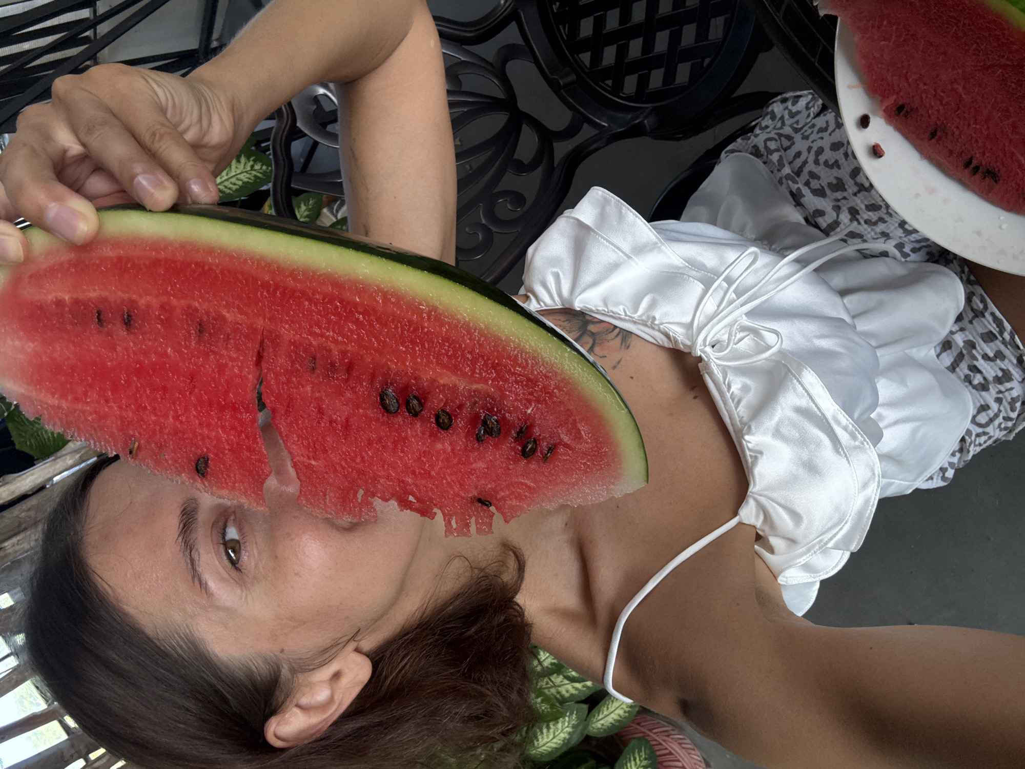 In the city of Nha Trang, Vietnam, a woman is pictured holding a slice of watermelon in front of her face. The woman has brown hair and is wearing a white ruffled top. Behind her, a black metal chair is visible, along with lush green plants. A plate with another slice of watermelon sits on a table to her right. The scene appears to be outdoors, possibly on a patio or balcony, given the natural lighting and the presence of plants and outdoor furniture.