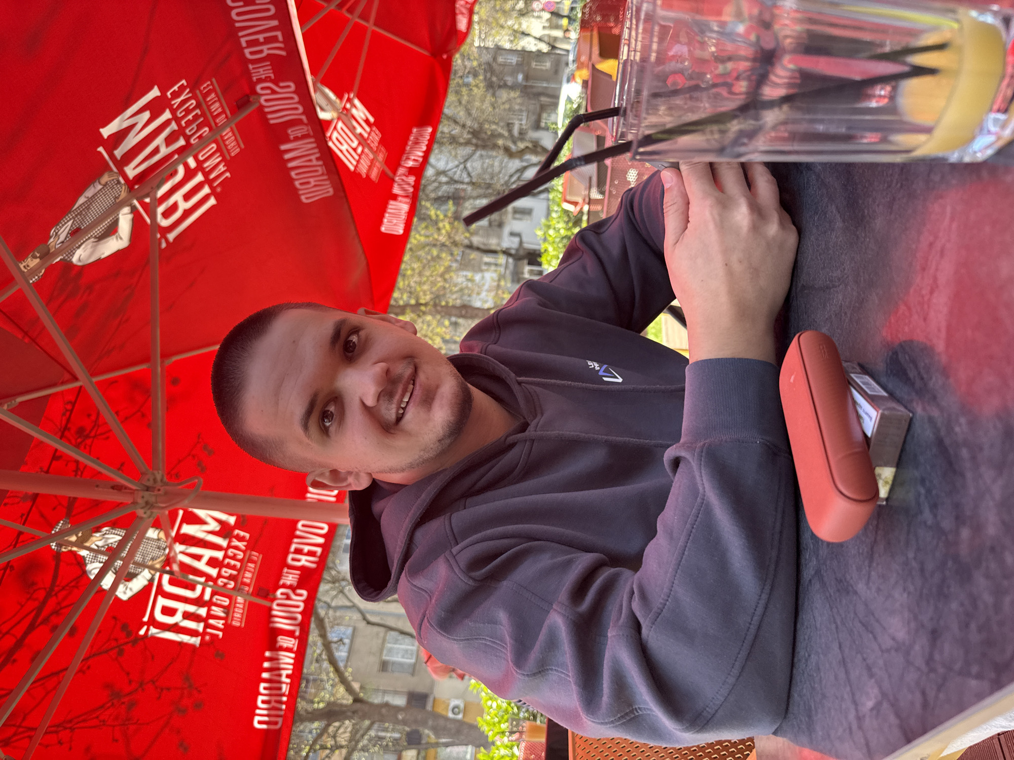 A medium, eye-level, angled shot of a man seated at an outdoor table. The man, who appears to be in his late 20s to early 30s, has short brown hair and is wearing a gray hooded sweatshirt. He is smiling slightly and looking directly at the camera. In front of him on the table is a tall glass with black straws, possibly containing a beverage with a slice of lemon. To his right is a red electronic cigarette case and a pack of cigarettes.
The scene is outdoors, under a large red umbrella with white text and designs. The text appears to be upside down and in reverse. Behind the man and the umbrella are trees with budding leaves, suggesting it is springtime. In the background, buildings are visible, indicating an urban setting.
Based on the text on the umbrella, it could be a branding of "WH5B", "ALAM", or "1804M". Due to this, a direct connection to Plovdiv, Bulgaria cannot be made.