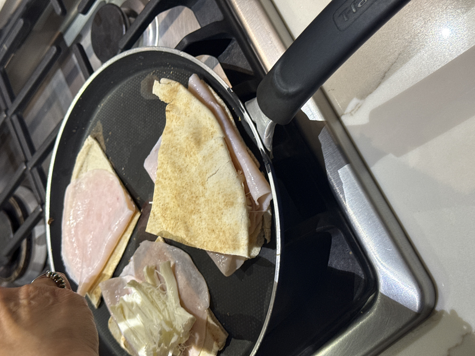 A close-up shot depicts a black frying pan on a gas stove, containing pieces of pita bread, sliced ham, and cheese. A hand wearing a ring is visible near the left edge of the pan. The pan is placed on the stove's burners. The background shows a countertop and a portion of the pan's handle. There is no evidence to determine if the location is San Bernardino Tlaxcalancingo, Mexico.