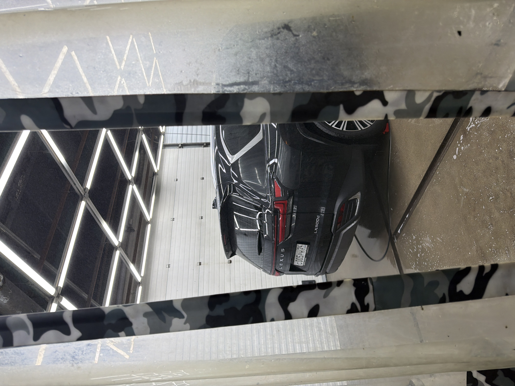 The image, taken from an elevated and obscured perspective, shows the rear of a black Lexus LX500d. The vehicle's license plate is partially visible, reading "C005PH 24," indicating the car is registered in the Krasnoyarsk region of Russia. The vehicle appears to be in a car wash facility, evidenced by the visible cleaning tools and spray. The lighting is bright and likely artificial. The foreground features a structure, possibly part of the car wash equipment, partially obscuring the view with a camouflage pattern design. The surrounding environment includes walls with fluorescent lighting and a textured surface.