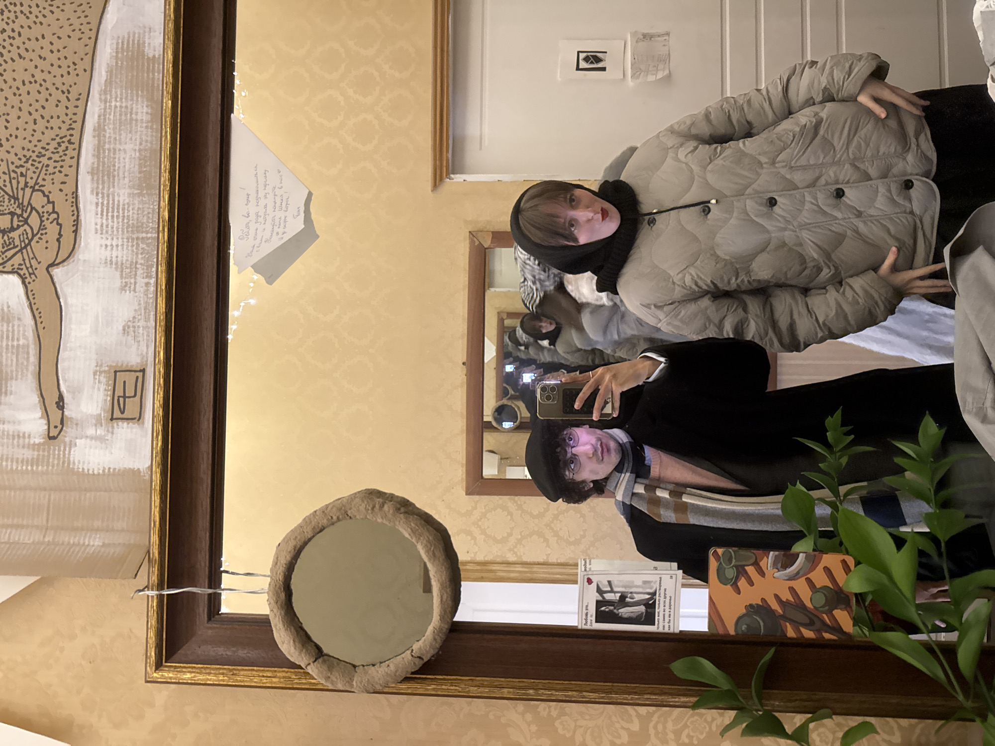 The scene shows a reflection in a mirror. Two people are visible, one taking a photo with a mobile phone and the other posing for the photo. The person taking the photo is wearing a black coat, beret, and striped scarf. The person posing is wearing a light gray padded jacket and a black scarf around their head.
The mirror has a wooden frame and several objects are visible on the shelf beneath it, including a round decorative mirror, a plant, and a small booklet. There is a piece of paper attached to the frame with writing in Russian. The wallpaper has a patterned design.
Based on the OCR text, it's possible the location is Moscow, Russia, as there's a note saying "Пете Шнка в мос".