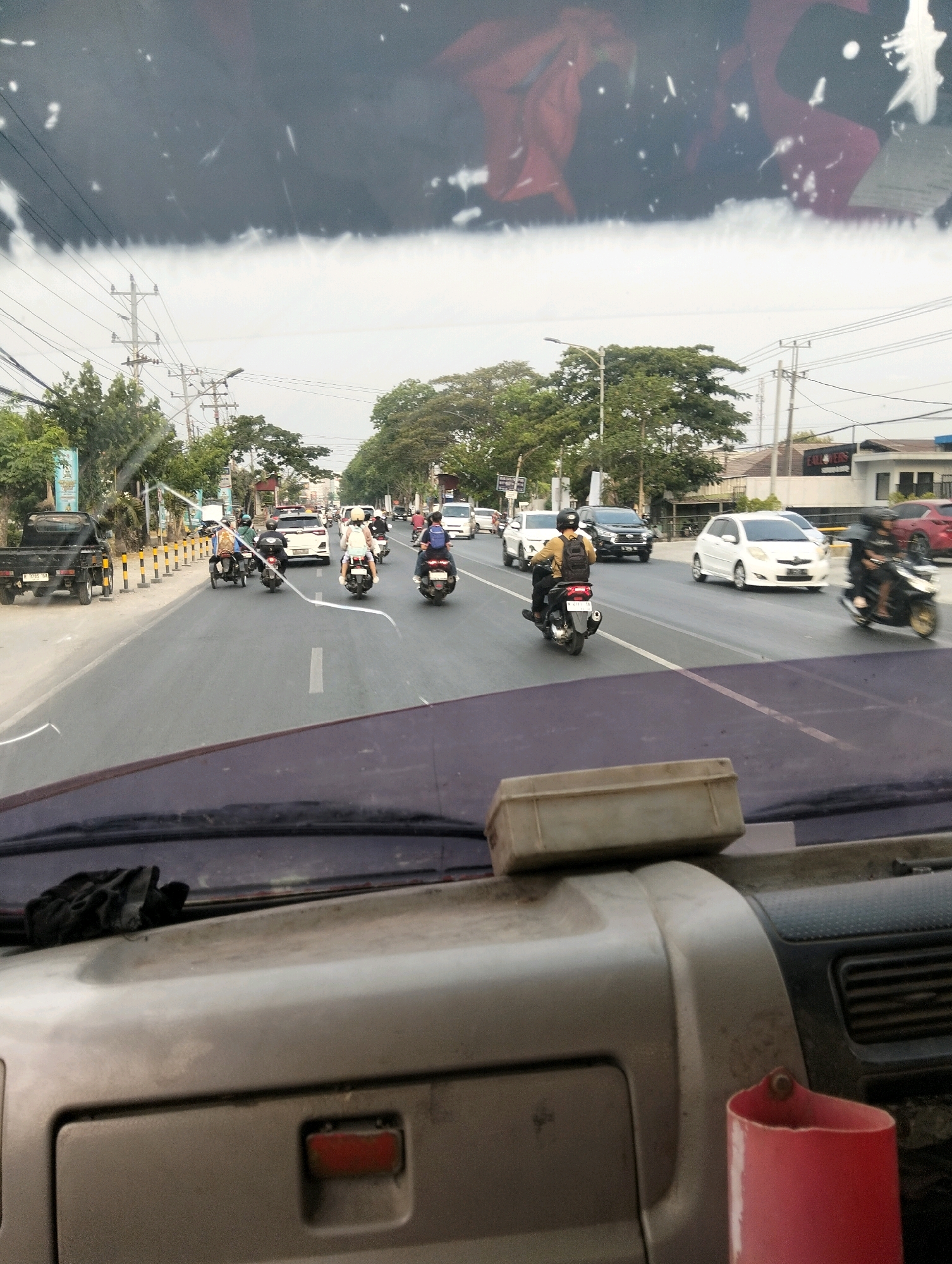The image depicts a view from inside a vehicle, looking out at a busy street scene in Semarang, Indonesia. The foreground shows the dashboard and windshield of the vehicle, with some smudges and glare on the glass. Beyond the windshield, traffic is visible on a multi-lane road. Several motorcycles are in motion, with riders wearing helmets and backpacks. There are also cars on the road, including a white sedan and a black SUV. Trees and utility poles line the side of the road, and buildings can be seen in the background. The overall impression is of a typical urban environment with moderate traffic flow. The presence of a black pickup truck with a license plate that appears to be Indonesian suggests the location is indeed Indonesia. The street signage is not clear enough to identify a specific street name, but the overall architectural style of the buildings and the types of vehicles are consistent with an urban setting in Southeast Asia.