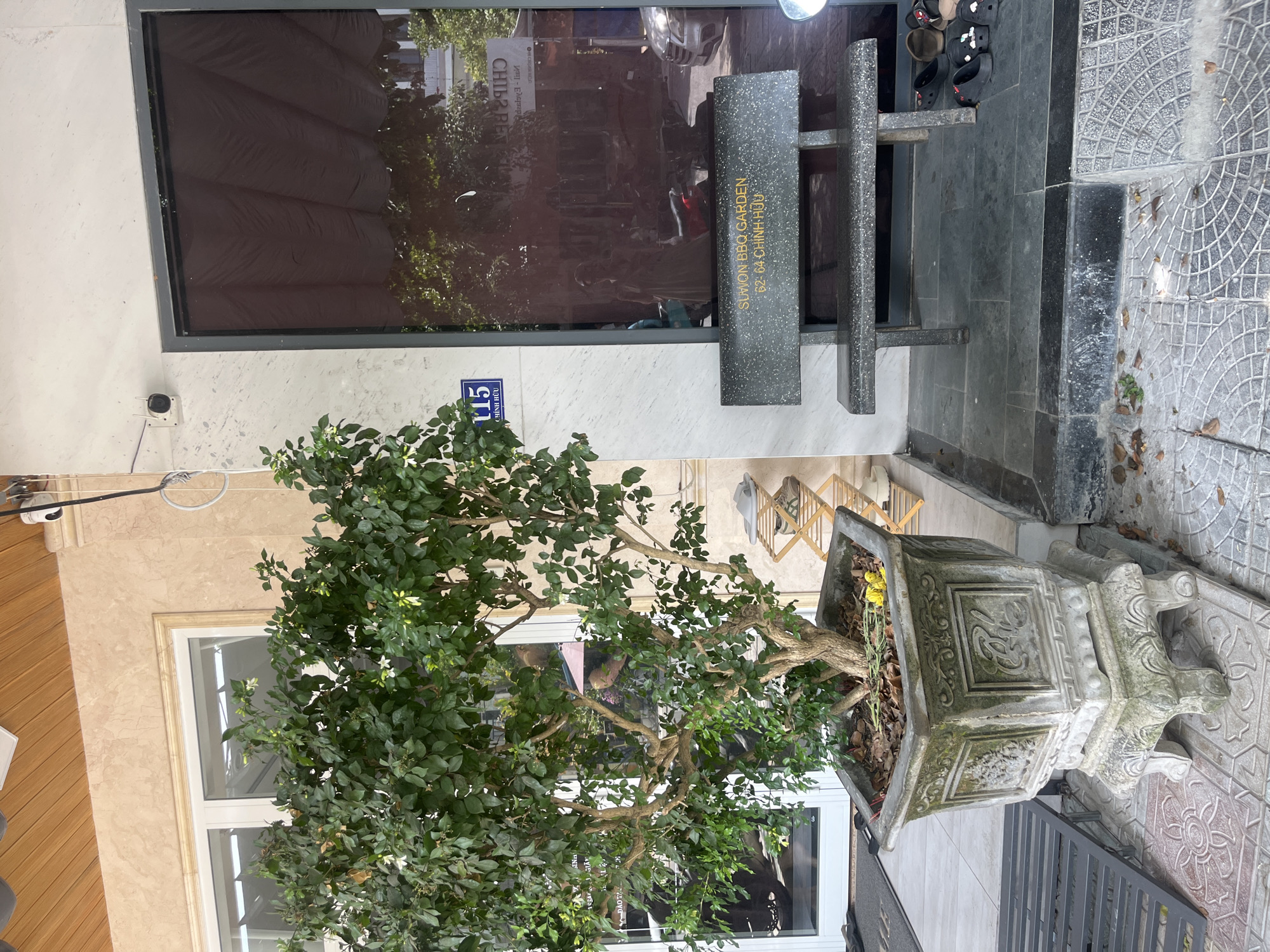 This image captures an outdoor scene in Đà Nẵng, Vietnam, likely in front of a commercial establishment. A potted tree with lush green leaves and small white flowers stands prominently in the foreground, housed in an ornate, weathered stone planter. To the right of the planter, a dark stone bench with the inscription "SUWON BBQ GARDEN 62-64 CHINH HUU" is positioned in front of a reflective glass door. The bench itself is speckled, suggesting it is made of granite or a similar composite material. A small wooden rack with shoes is visible to the right of the bench. The ground is tiled with a patterned paving. The building facade features light-colored marble-like cladding and a wooden ceiling detail. A security camera is mounted on the wall above the entrance. The reflection in the glass door shows a street scene with vehicles and a person.