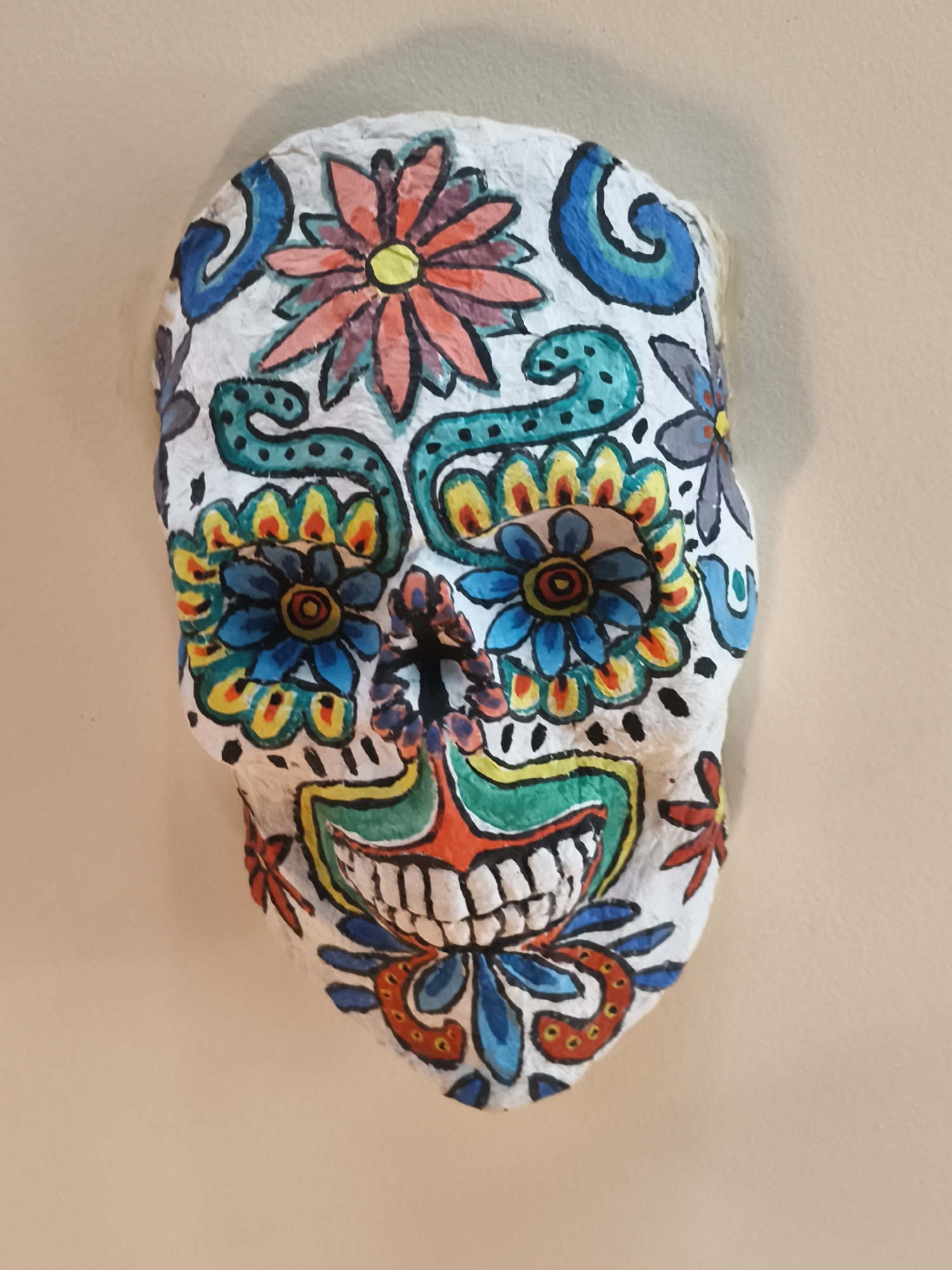 A single decorative skull mask is centrally presented against a plain, light beige wall. The mask, appearing to be crafted from a textured, possibly papier-mâché-like material, is predominantly white and adorned with vibrant, multicolored hand-painted patterns characteristic of a "sugar skull" or Day of the Dead aesthetic.
Detailed ornamentation includes:
- A large, multi-petaled orange-red flower with a yellow center at the top of the forehead.
- Two prominent blue floral patterns with orange and yellow centers represent the eye sockets, surrounded by teal, yellow, and red swirling motifs.
- A dark, irregular vertical slit forms the nose opening.
- Below the nose, a stylized mouth area features distinct white teeth against a red and green background, outlined in black.
- Additional blue, teal, red, yellow, and gray floral and swirling designs populate the cheeks, chin, and sides of the mask. All patterns are outlined in black.
The mask is illuminated, casting a soft shadow on the wall behind and to its upper right, suggesting an indoor environment. No individuals are present, and no actions or interactions are depicted. The scene is located in Varna, Bulgaria.