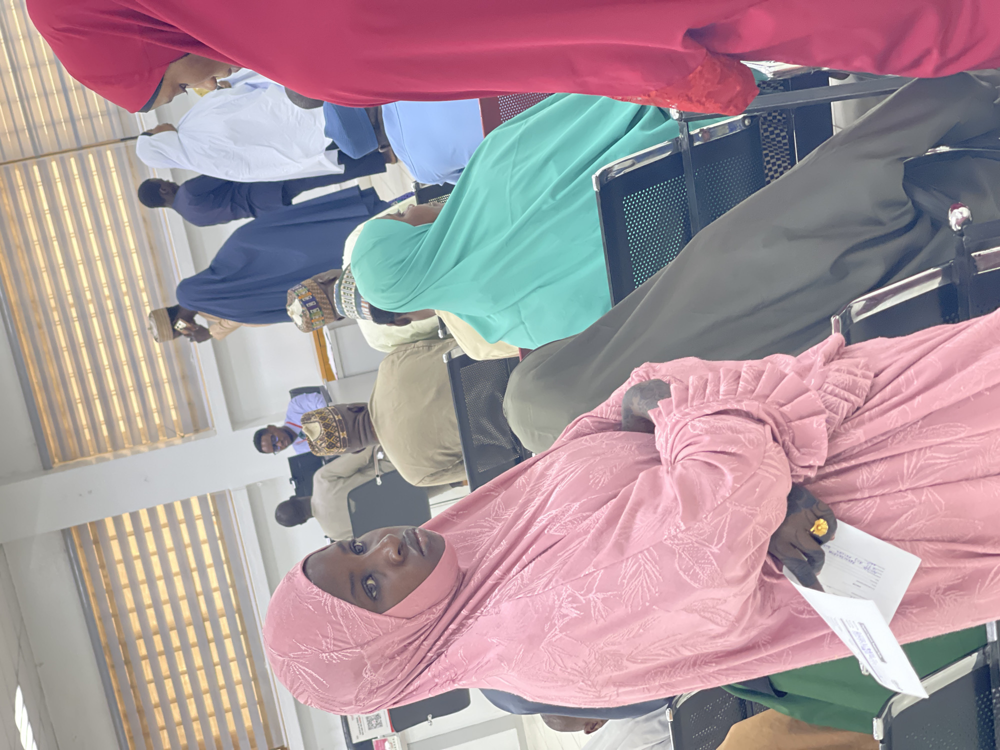 The image captures an indoor gathering of individuals, predominantly adults, seated in rows of chairs, rotated 90 degrees clockwise. The setting appears to be a classroom, lecture hall, or conference room with light-colored walls and windows featuring vertical blinds in the background, suggesting bright daytime lighting.
In the immediate foreground, a woman with dark skin wears a pink hijab and a patterned long-sleeved dress with ruffled cuffs. She looks calmly towards the camera, holding several white papers in her right hand, which also displays a gold-colored ring. Behind her, other attendees are visible. To her left, another individual wears a teal or mint green hijab and dress. Further back, men and women are dressed in various traditional and modest attire, including white thobes, navy blue robes, light blue shirts, and traditional patterned caps (kufi). The chairs are black with metal frames, consistent with institutional seating.
The people are seated in an organized manner, facing forward, indicating they are attending a lecture, meeting, or training session. The papers held by the woman in pink could be notes, registration forms, or event materials. The style of dress, including hijabs and kufi, provides a strong cultural cue consistent with the provided location context of Zaria, Nigeria. A partially visible QR code is present on a sign or sticker in the lower left corner of the image, but its content is illegible. No other text on the papers or elsewhere is clearly readable. The atmosphere appears focused and orderly.