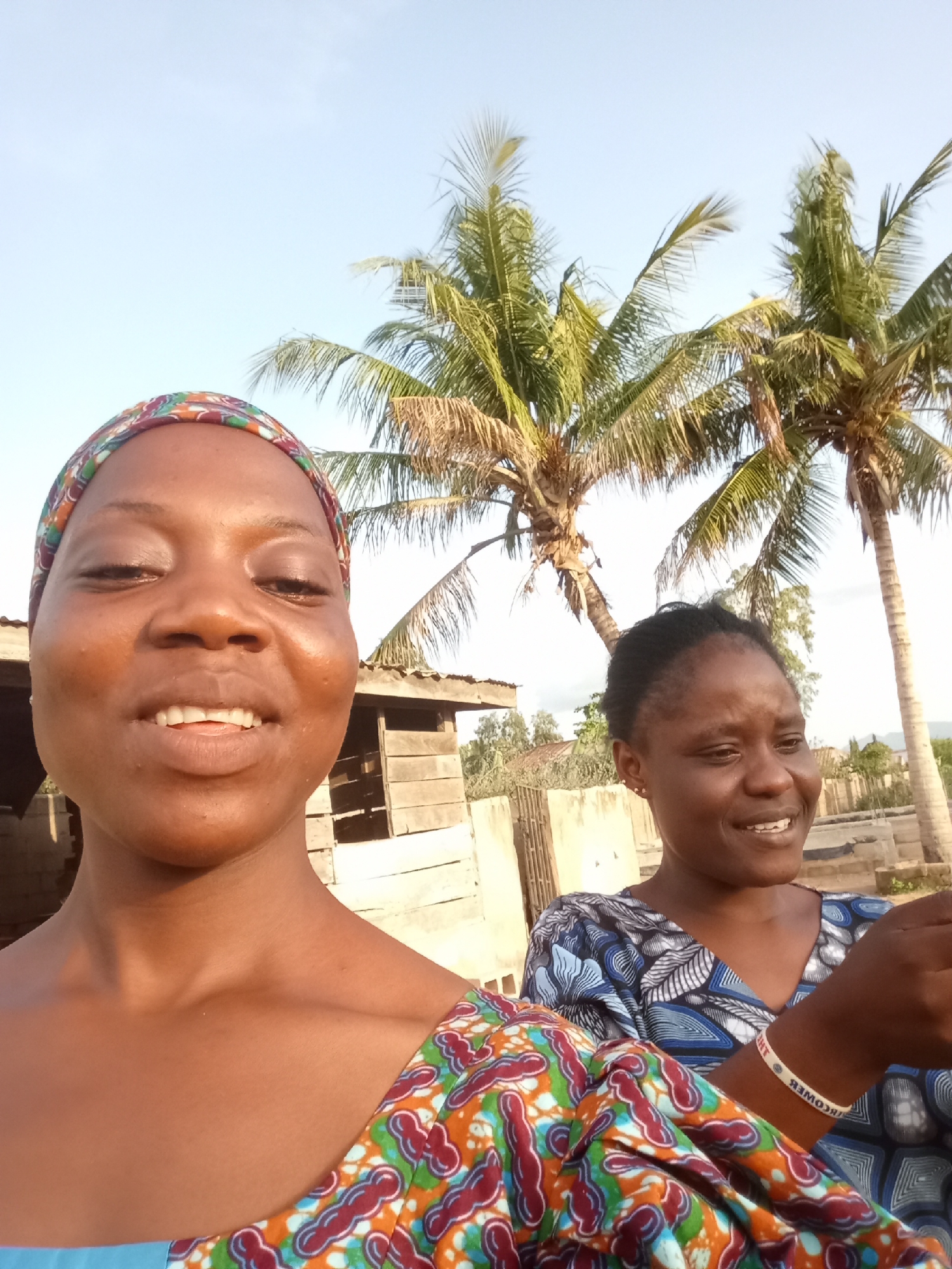 This media file shows a close-up selfie of two Black women outdoors in a tropical or sub-tropical setting, likely Jalingo, Nigeria. The woman in the foreground is closer to the camera, facing it with a slight smile and her mouth open. She is wearing a colorful headwrap with patterns of red, green, orange, and blue, and a matching garment on her shoulder. Her skin is brown, and she has dark eyes and hair.
Behind her, the second woman is visible, also looking towards the right and smiling. She is wearing a blue and white patterned top. Her hair is pulled back. A thin white bracelet with black text is visible on her right wrist.
The background features elements of a rural or semi-rural environment. Two palm trees are prominent, with their fronds spread against a clear, pale blue sky. To the left of the women, there is a rustic wooden structure, possibly a shed or a simple building with wooden planks. Further in the background, there are more structures and fences, suggesting a village or settlement. The lighting suggests it is daytime, likely late afternoon or early morning due to the warm tones and long shadows. The overall mood appears cheerful and relaxed. The text on the bracelet appears to read "NEVER GIVE UP" in reverse.