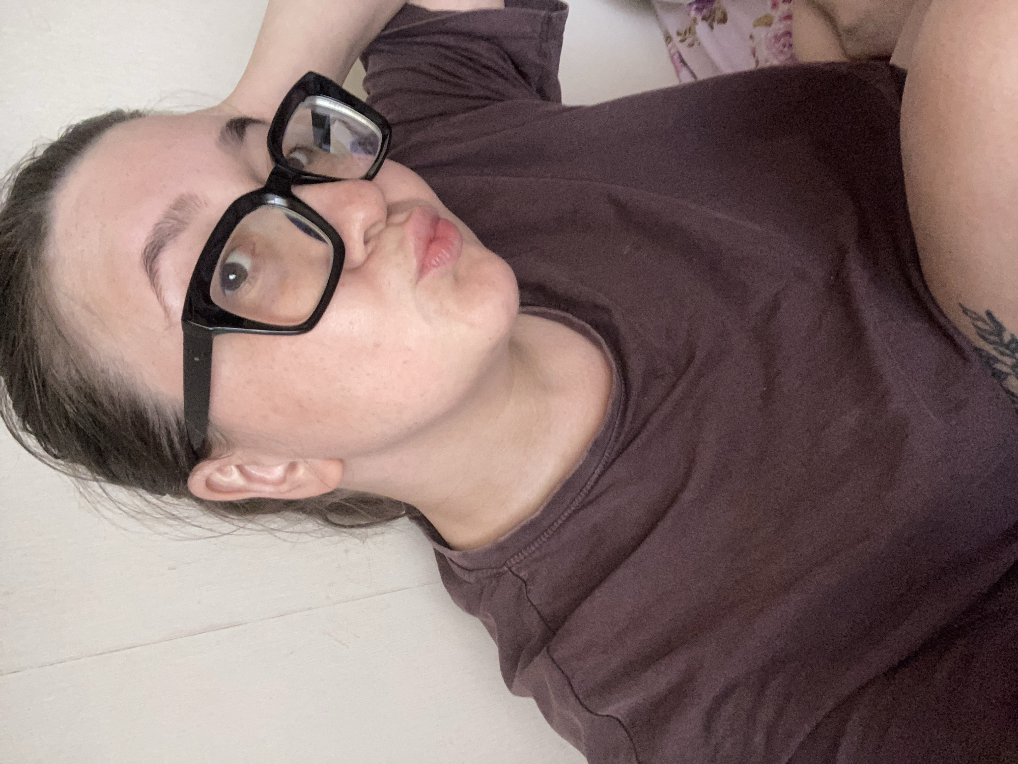 A woman is lying on a white floor, wearing glasses with black frames and a brown t-shirt. She is looking up and slightly to the left with her lips puckered. Her head is resting on her hand, which is positioned behind her head. A small tattoo is visible on her upper arm. The background is blurred, but a floral patterned blanket is partially visible. The location cannot be determined from the image.