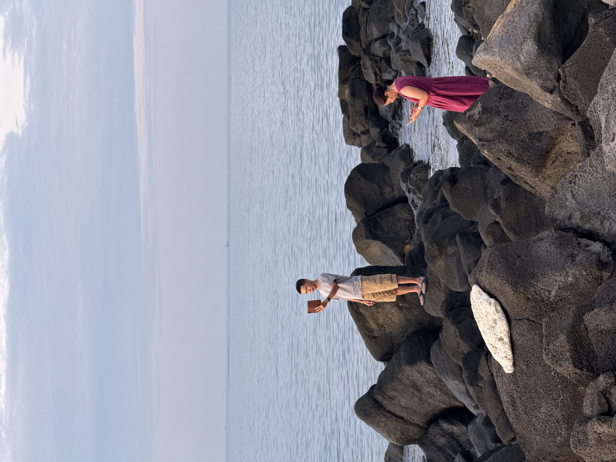 Two individuals are on a rocky shoreline at sunset. A man in a gray t-shirt and khaki shorts stands on dark, jagged rocks, holding a tablet computer in his right hand, appearing to take a photo of himself. He is barefoot, wearing flip-flops. To the right, a woman in a magenta dress stands on similar rocks, looking down at her smartphone. She has her hair pulled back. The sea is calm and stretches to the horizon under a pale, cloudy sky. The location is identified as Lý Sơn Special Administrative Region, Vietnam.
