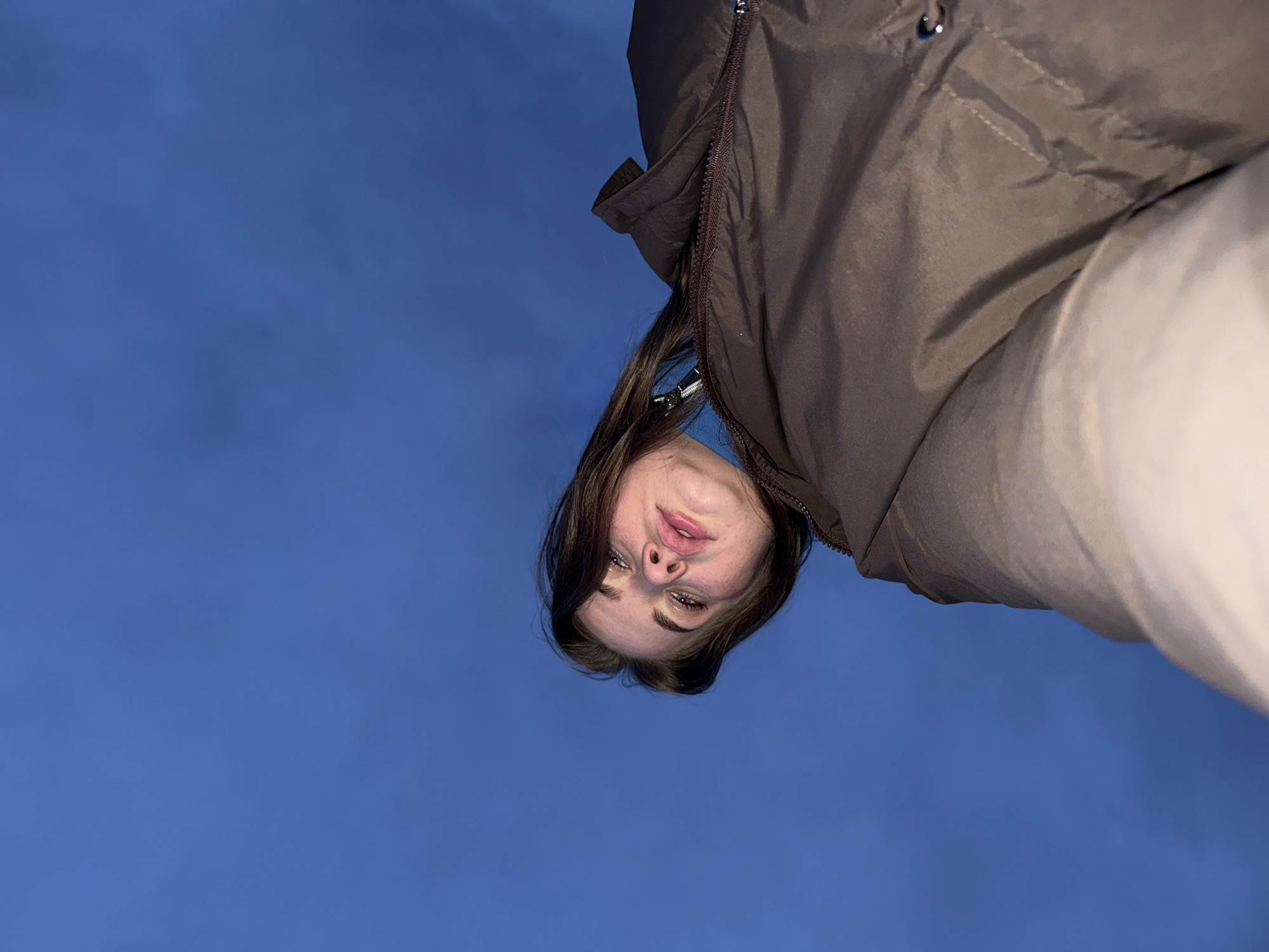 An unusual, low-angle portrait captures a young woman positioned upside down against a deep blue sky in Ol'khovochka, Russia. Her head and upper torso are visible in the upper part of the frame, with her gaze directed downwards towards the viewer. She has dark brown hair, and her eyes are open, focused on the camera. Her expression is neutral to slightly contemplative, with a subtle pout to her lips. She is wearing a dark brown puffer jacket, part of which is visible, featuring a prominent zipper along the front opening. A lighter, possibly off-white or beige, garment is also visible on the right side of the frame. The background is an expansive, uniform dark blue sky, suggesting either a twilight setting (dusk or dawn) or a clear, cloudless day with a deeply saturated blue hue. No other objects or text are visible in the image.