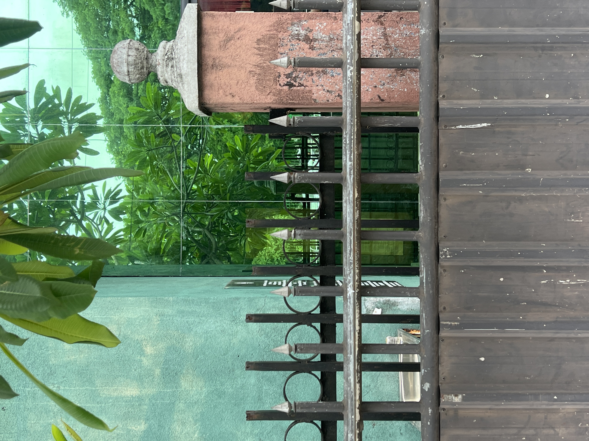 A close-up, eye-level outdoor shot captures a metal gate and fence in front of a textured teal wall. In the background, lush green trees are visible through a reflective glass surface, creating a mirrored effect. The fence is made of vertical metal bars with pointed tips, interspersed with circular elements. A brick pillar, painted a faded reddish-brown with visible wear and tear, supports the fence on the right side, topped with a decorative stone finial. The gate itself is a solid, dark metal surface with vertical corrugated panels. Leaves from a plant hang into the frame from the top left. The overall scene suggests an urban environment with greenery and architectural elements.