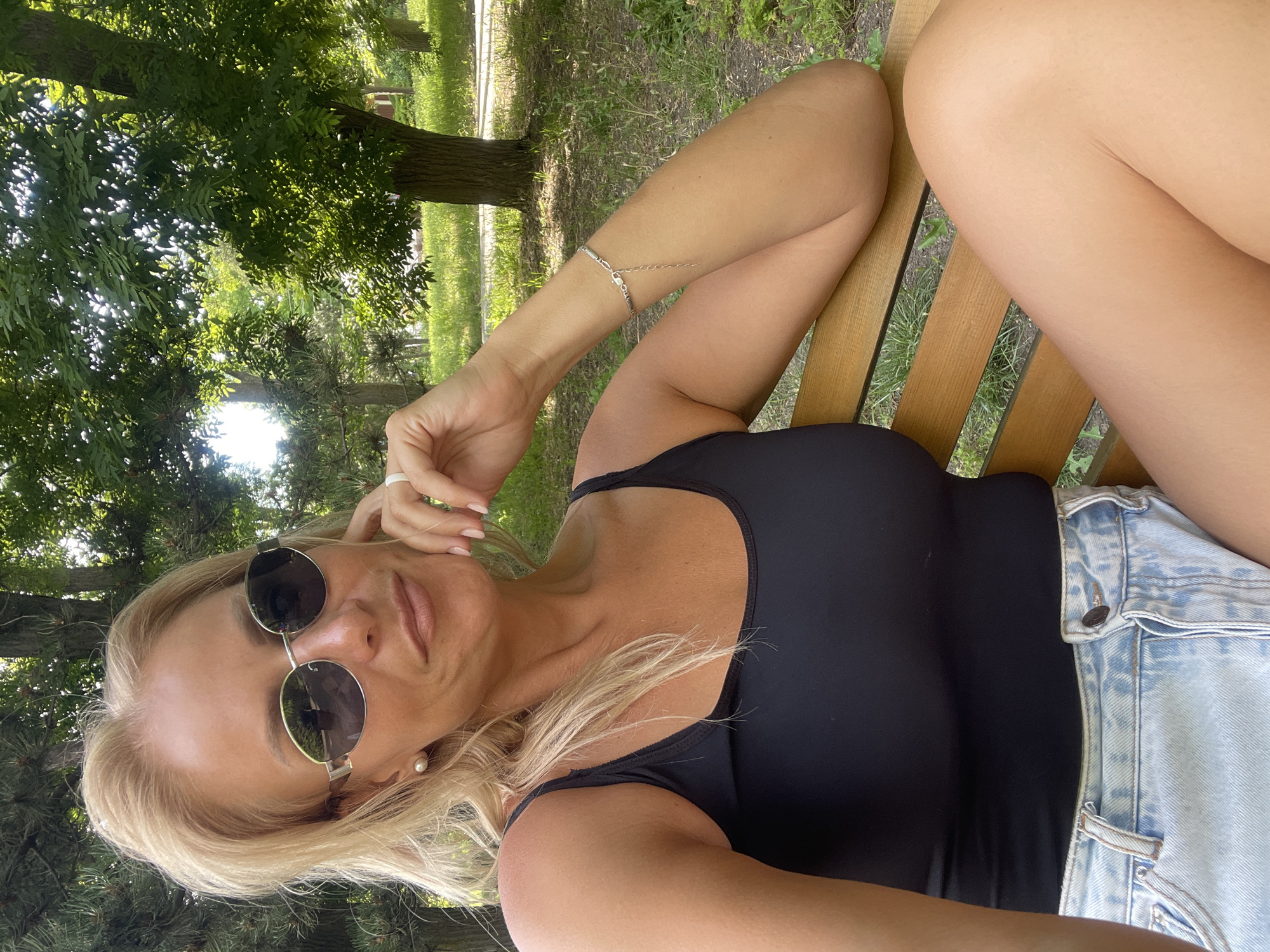 A woman with blonde hair and sunglasses is lying on a wooden bench in a park. She is wearing a black tank top and denim shorts. The sun is shining through the trees, casting dappled shadows on the grass. The woman is resting her head on her hand and appears relaxed. The overall mood is peaceful and serene. The time of day is likely daytime, given the bright sunlight. There is no visible text in the image.