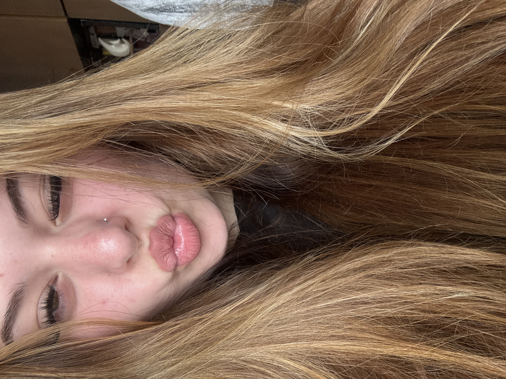 The image is a close-up portrait, rotated 90 degrees counter-clockwise, featuring a young person with long hair. The subject's face is partially visible, primarily focusing on her nose and mouth, along with her left eye and eyebrow. Her lips are pursed in a "kissing" or "duck face" expression, enhanced with a light pink, possibly glossy, lip product. A small, shiny stud nose piercing is prominent on her right nostril. Her eyes are partially closed, revealing dark, possibly mascara-coated, eyelashes. Her fair skin shows a slight redness on the bridge of her nose and cheeks.
The most dominant feature in the image is her long, flowing hair, which occupies a large portion of the frame. The hair is a blend of light brown and blonde tones, with visible highlights, appearing soft, slightly wavy, and well-maintained. It cascades around her face, almost framing it.
The background is largely obscured by her hair, but indistinct dark and brown shapes, along with a faint white object, are visible in the upper left corner, suggesting an indoor setting. There is no discernible activity other than posing for the photo. The overall impression is casual and playful. No text is visible within the image. The provided location context is Troyan, Bulgaria.